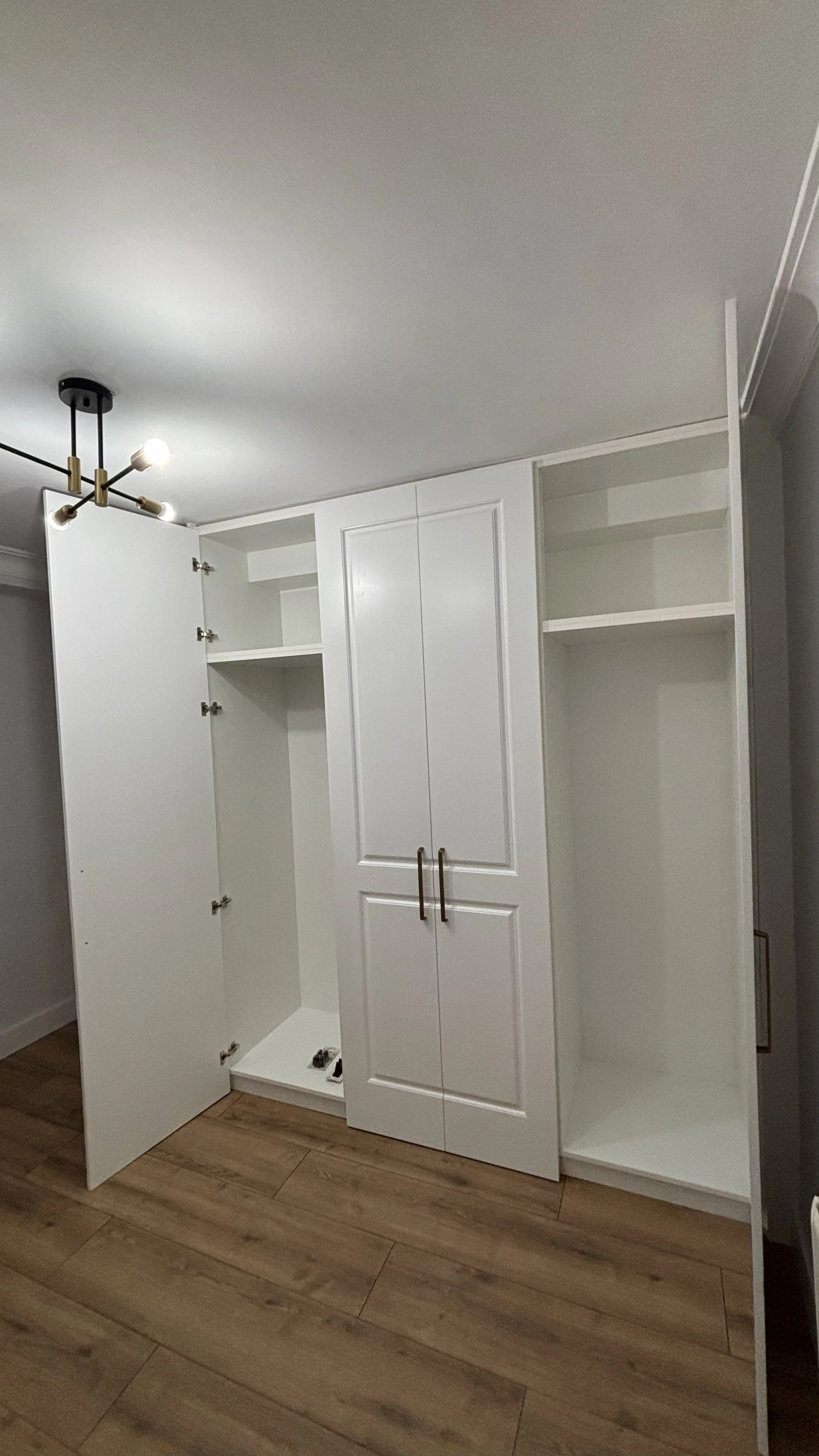 Armario blanco en proceso de instalación con puertas abiertas y estantes internos, instalado en una habitación con piso de madera y luces en el techo.