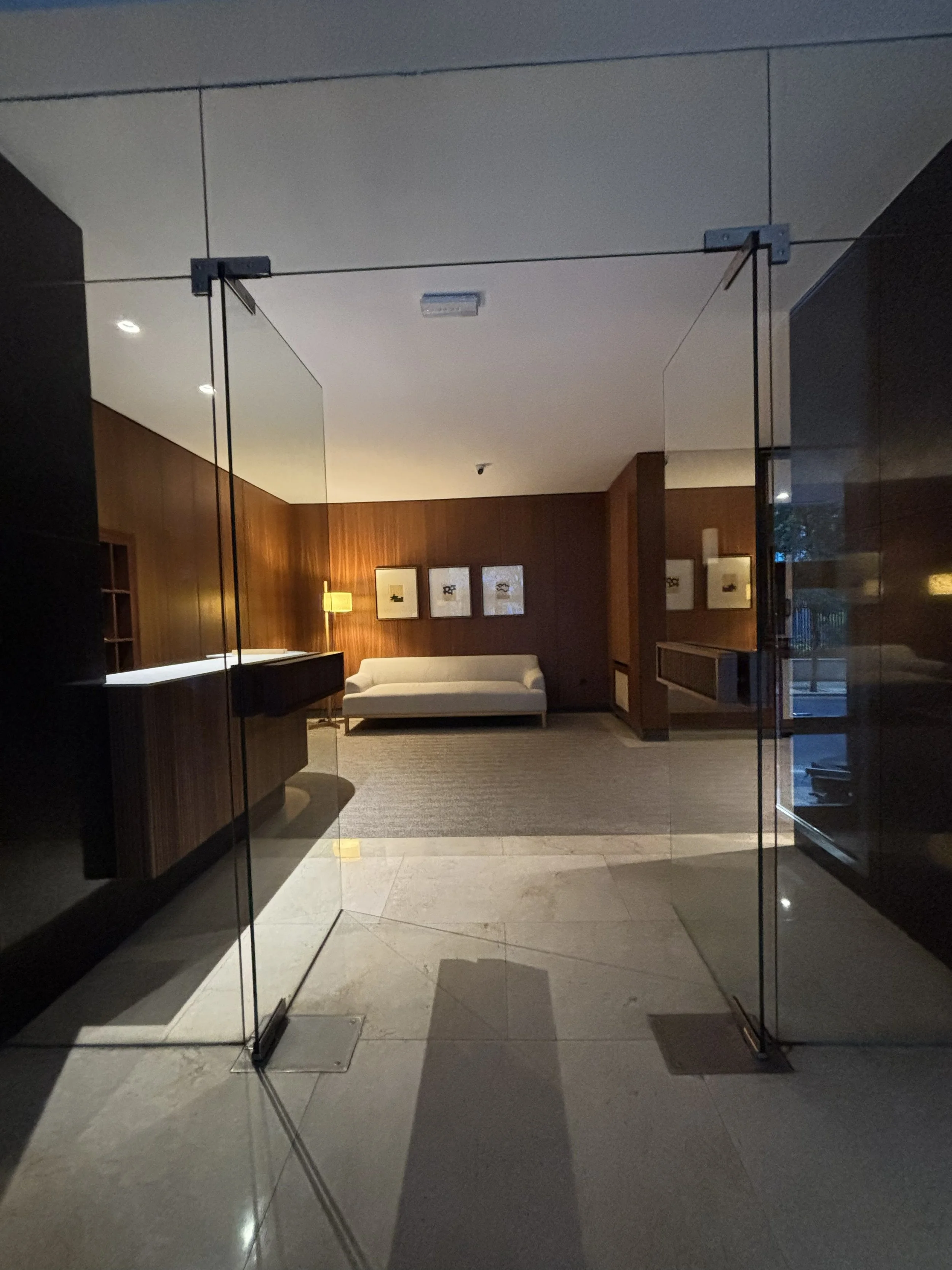 Vestíbulo con puerta de vidrio y sala de estar con sofá beige y cuadros en la pared de madera.