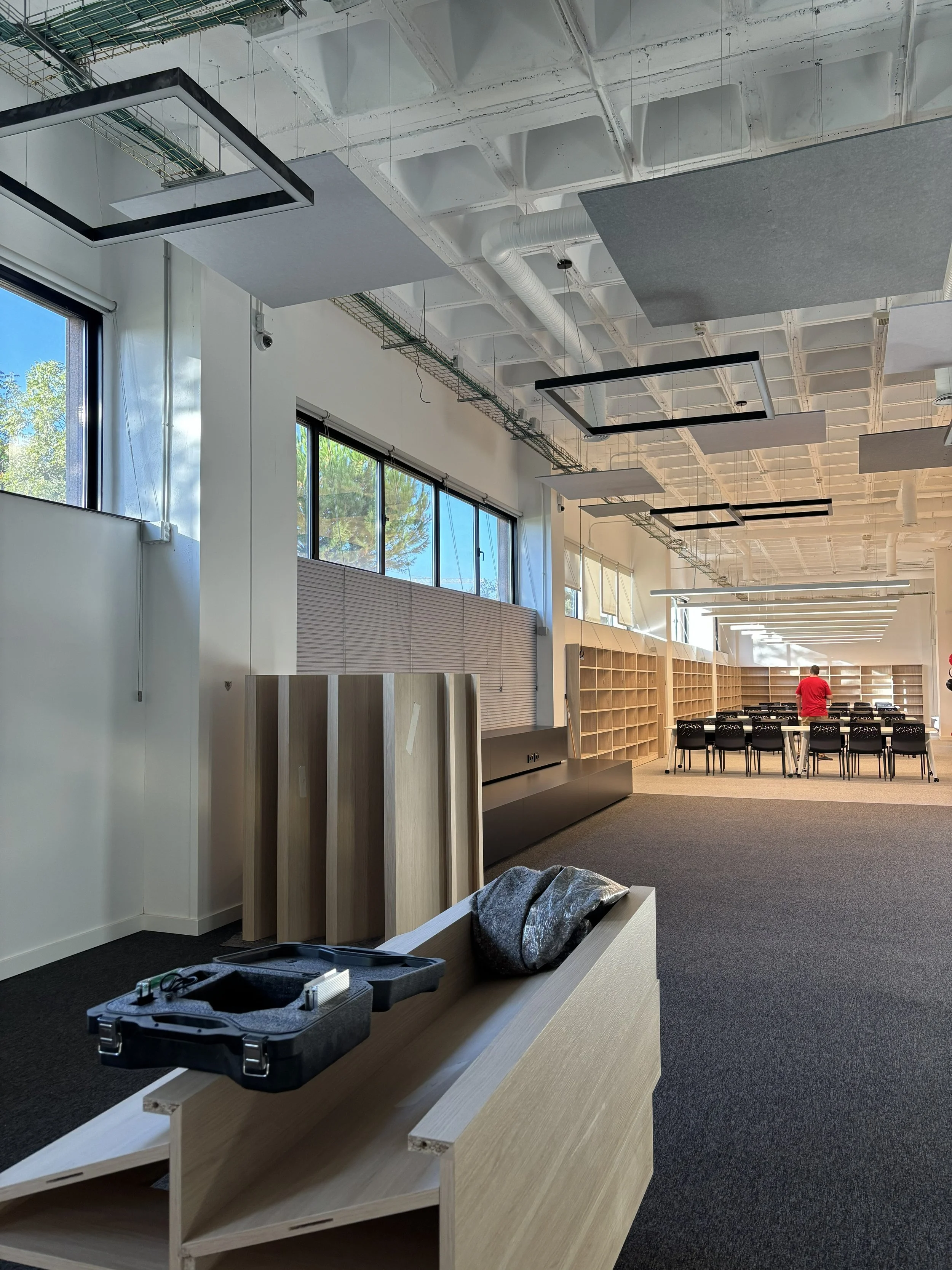 Espacio interior en proceso de amueblamiento con estanterías de madera, mesas y sillas, ventanas grandes que dejan entrar luz natural, y un techo con paneles acústicos y sistemas de ventilación.