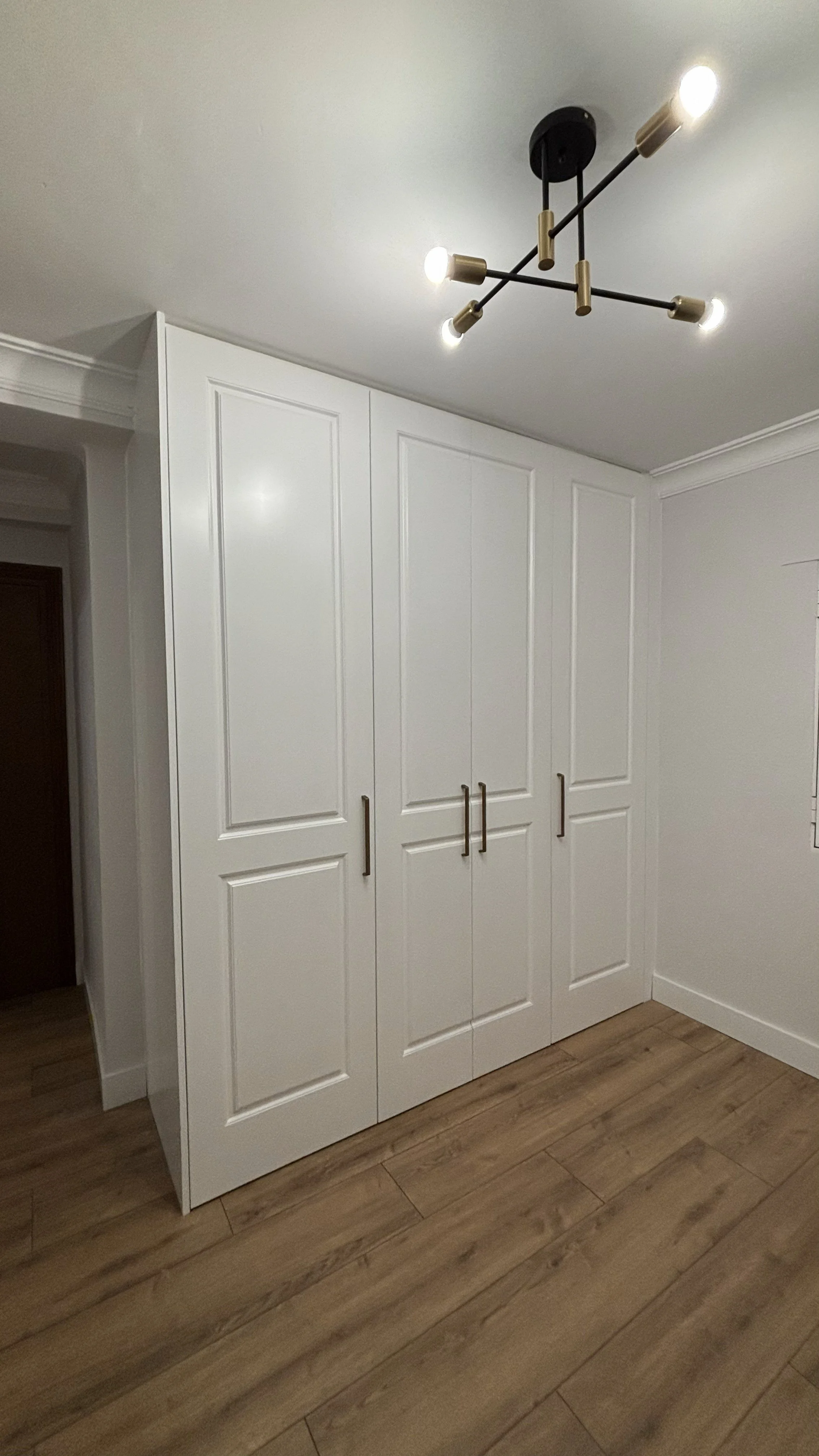 Vista de un armario blanco con puertas de panel y asas doradas en una habitación con piso de madera y techo con lámpara moderna de bombillas expuestas.