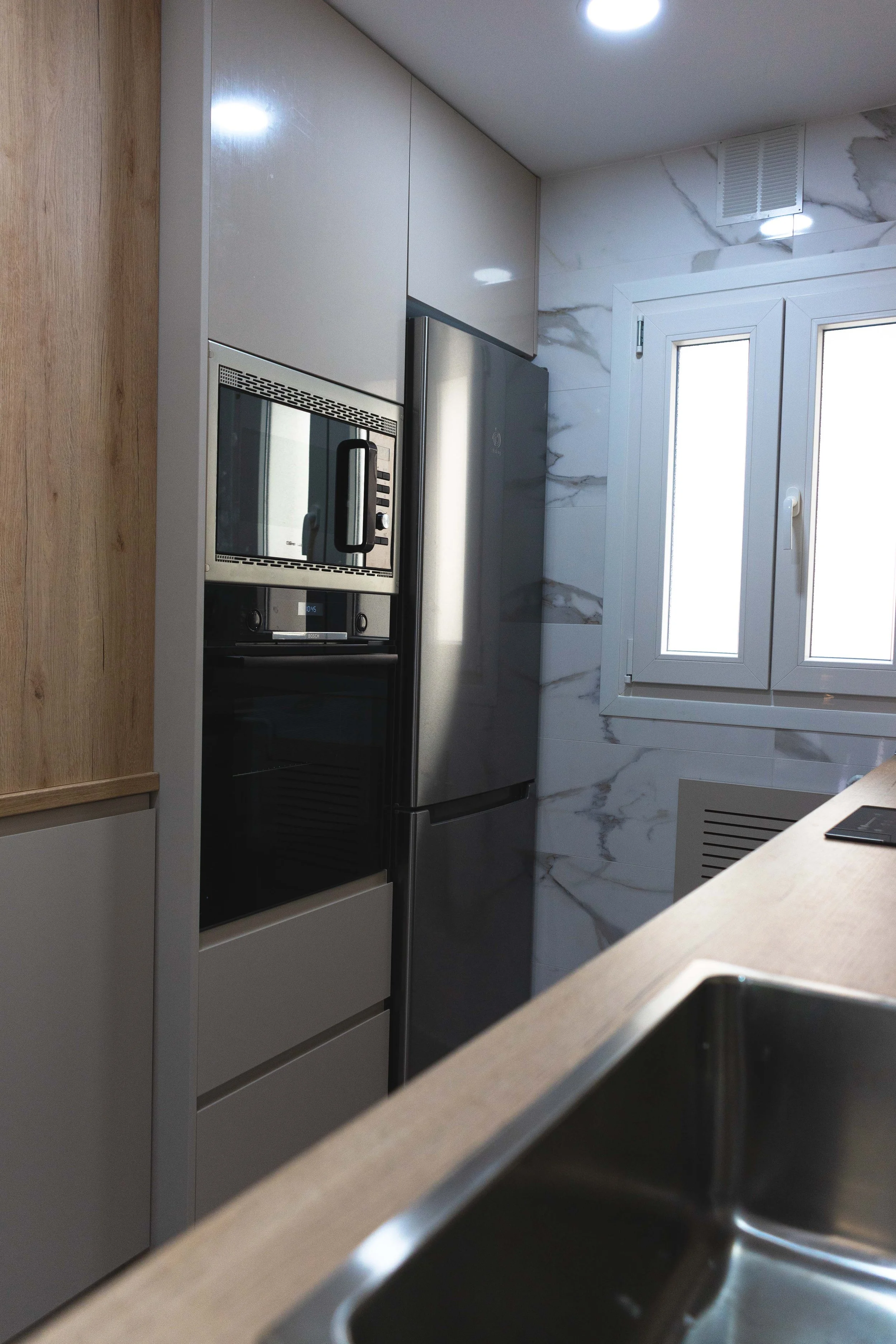 Cocina moderna con electrodomésticos de acero inoxidable, ventana con persiana, paredes de mármol y encimera de madera.