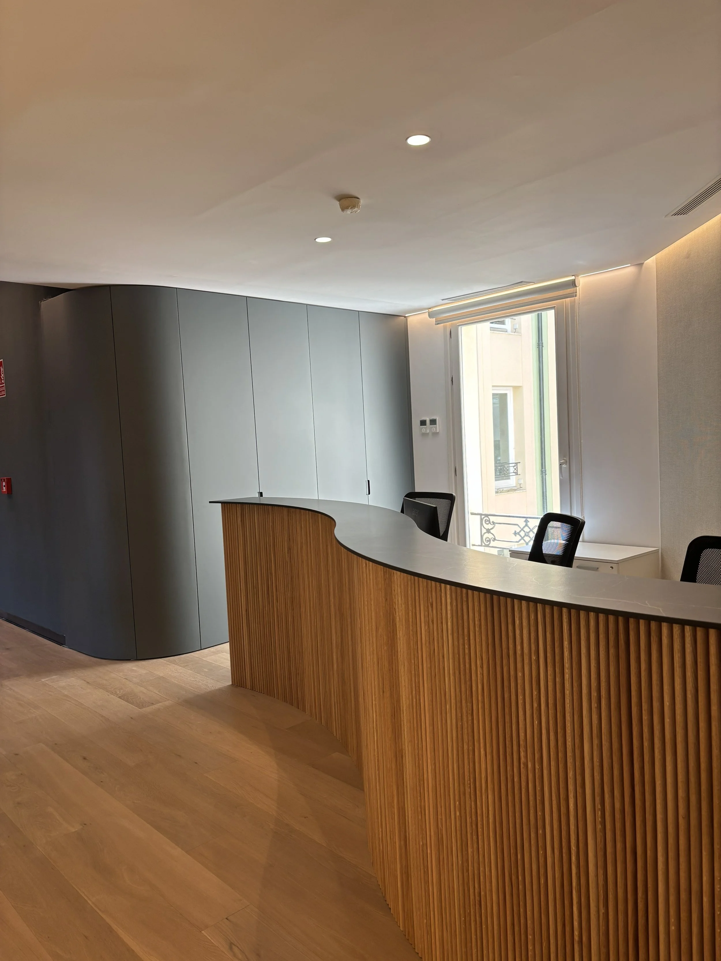 Mostrador de madera en una recepción moderna con sillas negras, ventana y paredes blancas.