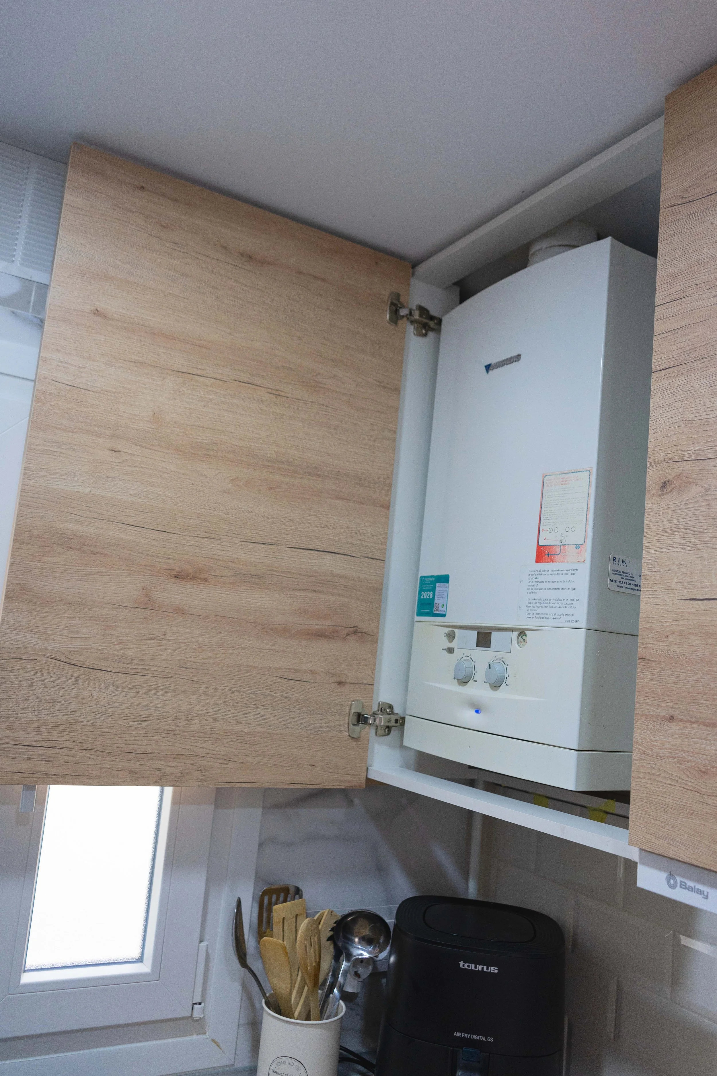 Estufa de agua calentan en un armario de cocina con utensilios de madera y electrodoméstico cerca de una ventana.