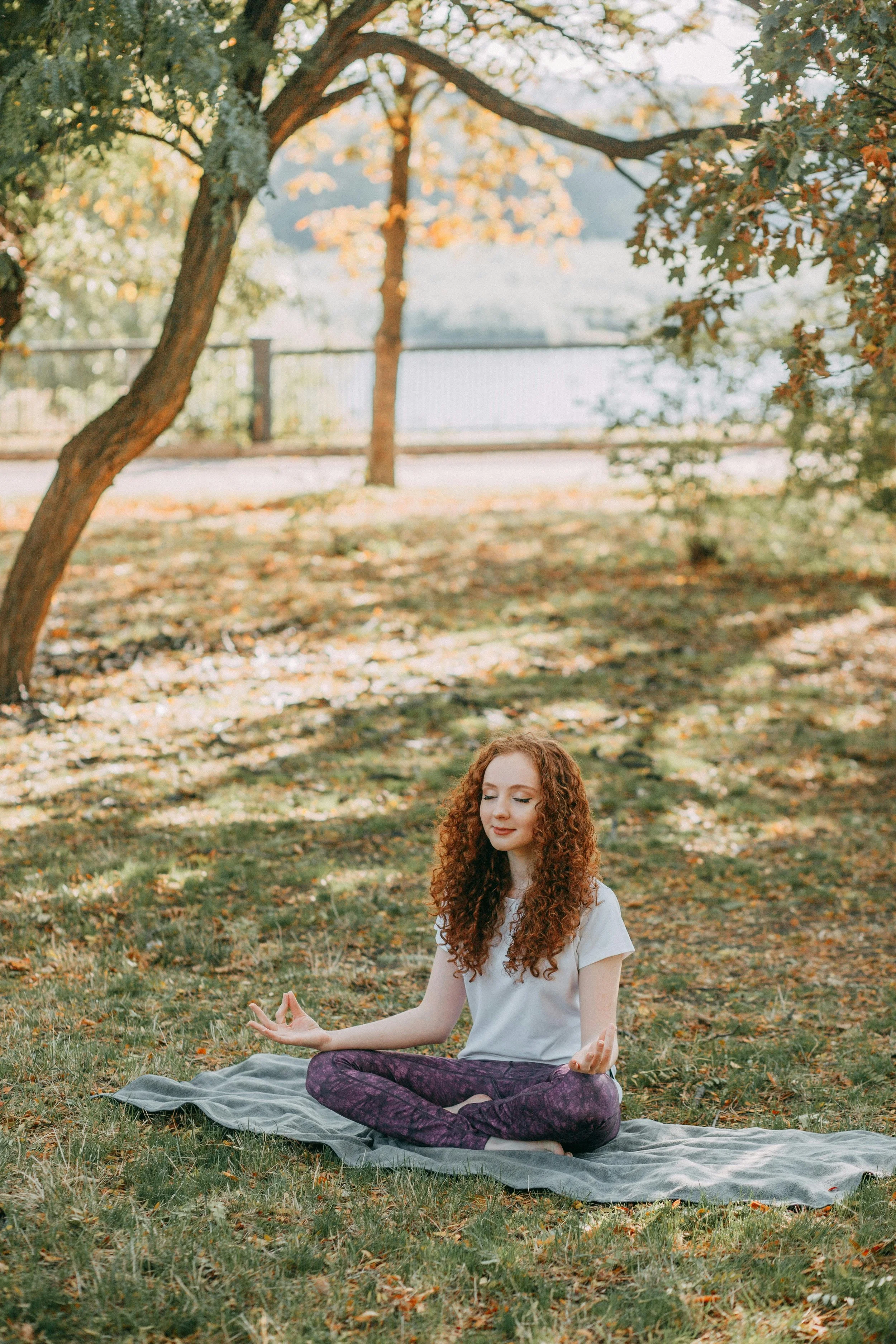 An image depicting a young woman with curly red hair practicing yoga outdoors on a mat in a park with autumn leaves and trees representing therapy for women and therapy for college students specializing in Anxiety Therapy in Lansing, MI.