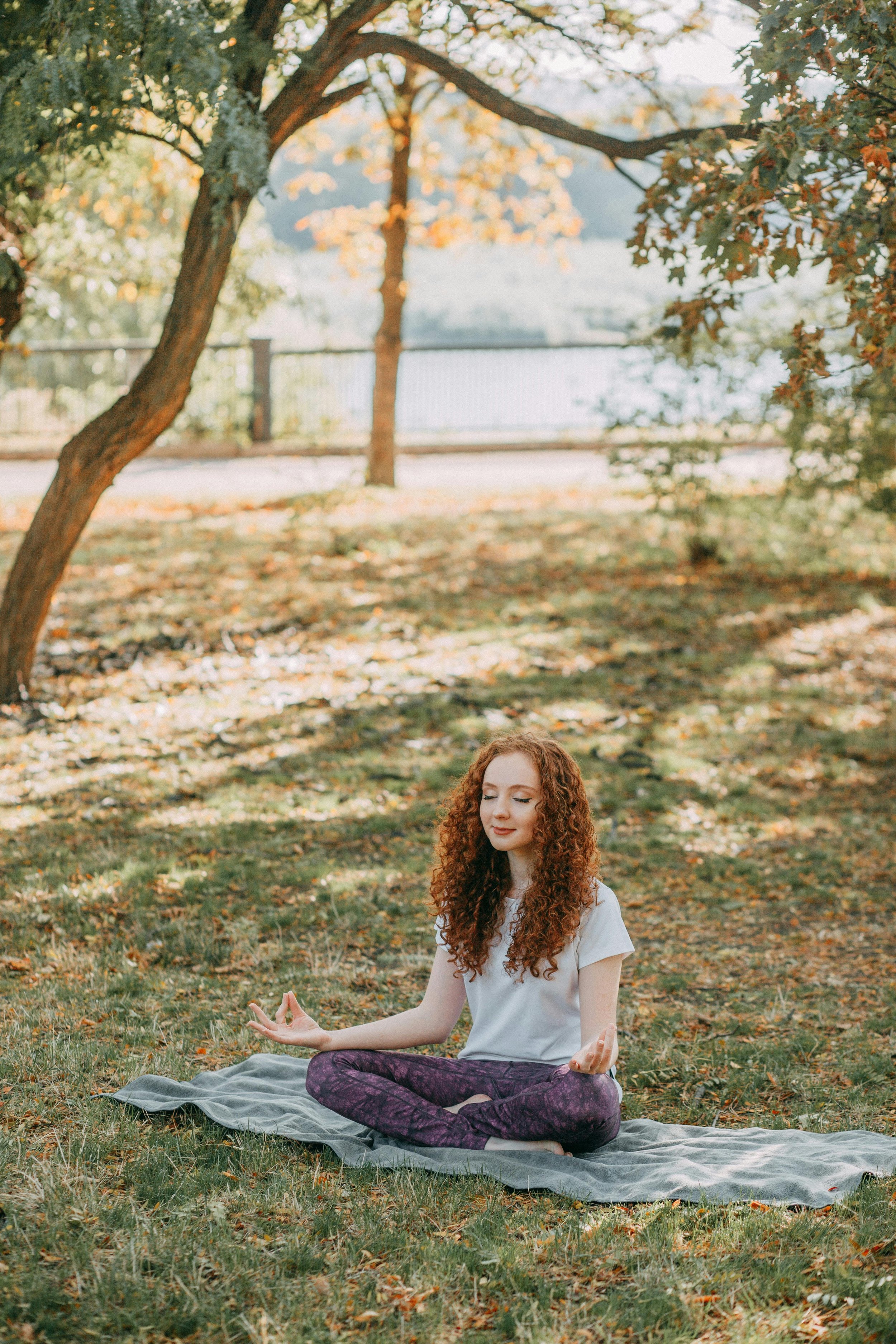 An image depicting a young woman with red curly hair practicing yoga outdoors in a park, sitting cross-legged on a mat representing therapy for women and therapy for college students specializing in Anxiety Therapy in East Lansing, MI.