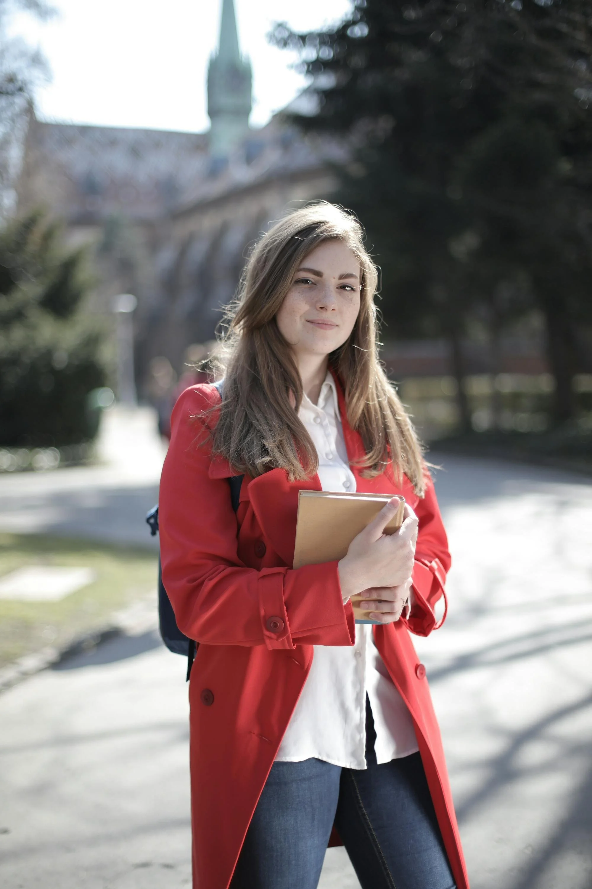 A young woman stood outdoors on a sunny day, holding a notebook, wearing a red coat and jeans with a building and trees in the background.