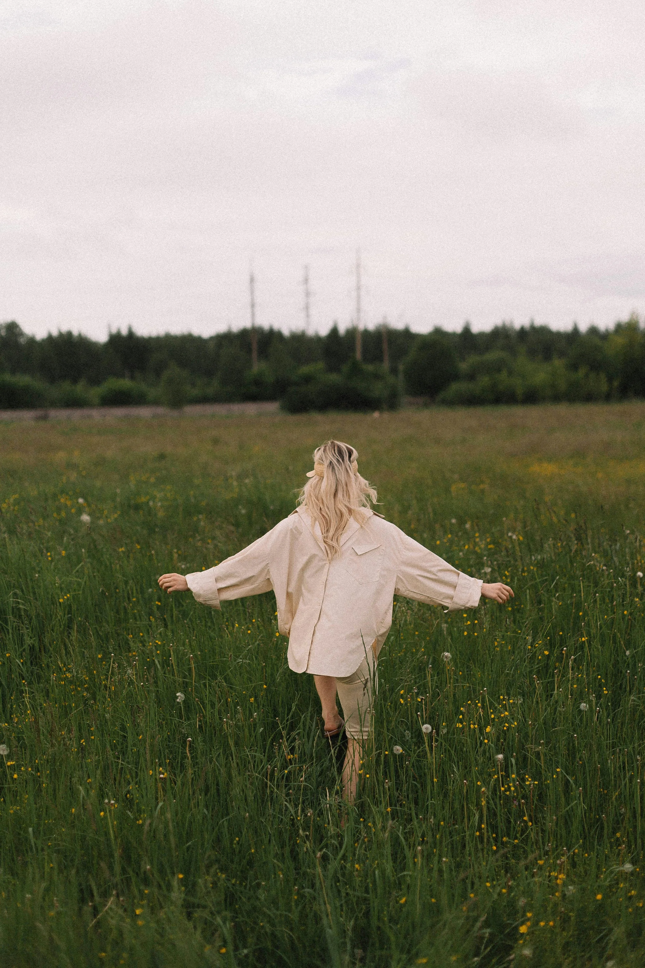A woman in a beige coat walking barefoot through a grassy field with yellow flowers, facing away from the camera, with trees and power lines in the background under a cloudy sky.
