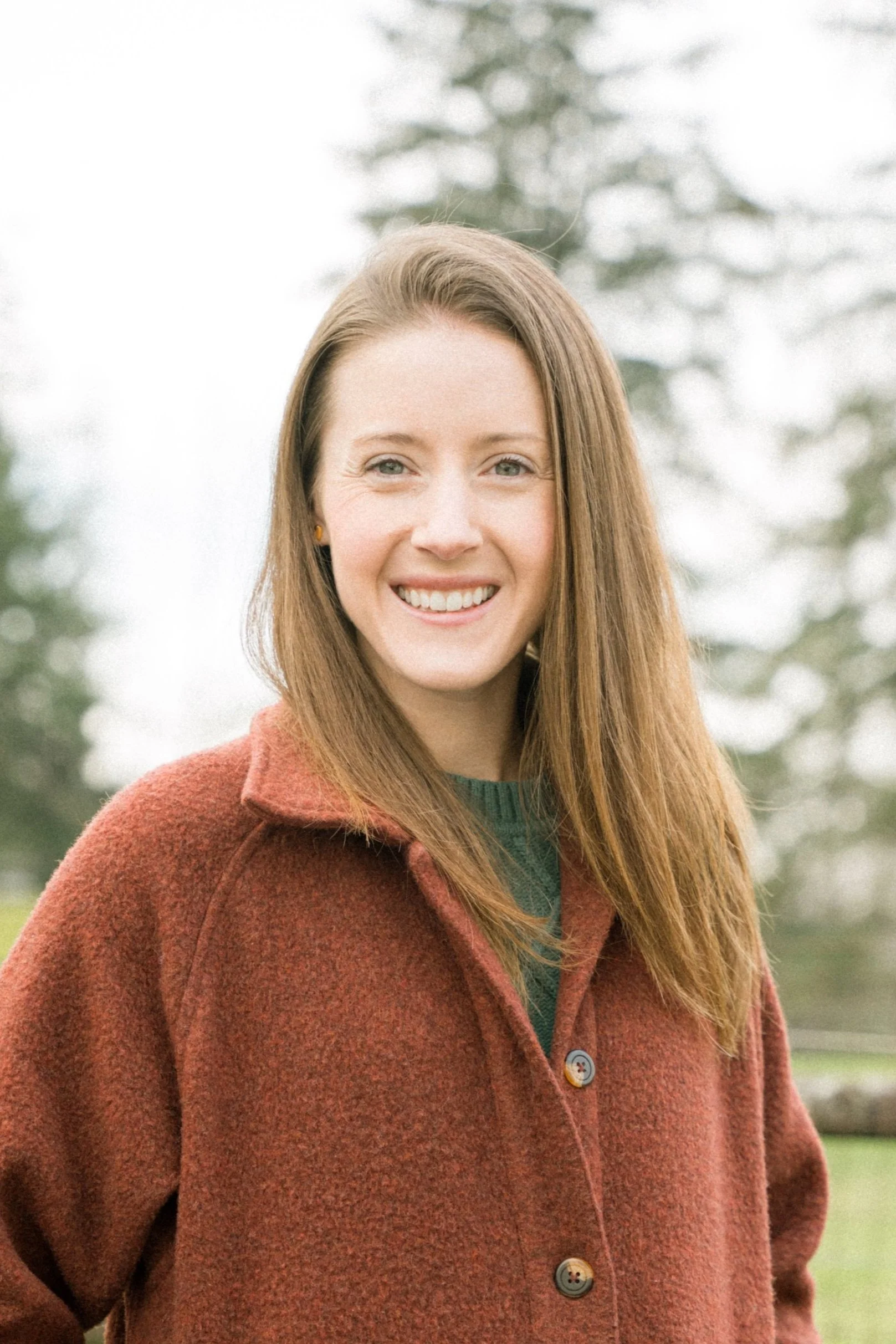 A smiling young woman with long red hair, wearing a rust-colored fleece jacket outdoors with trees in the background.