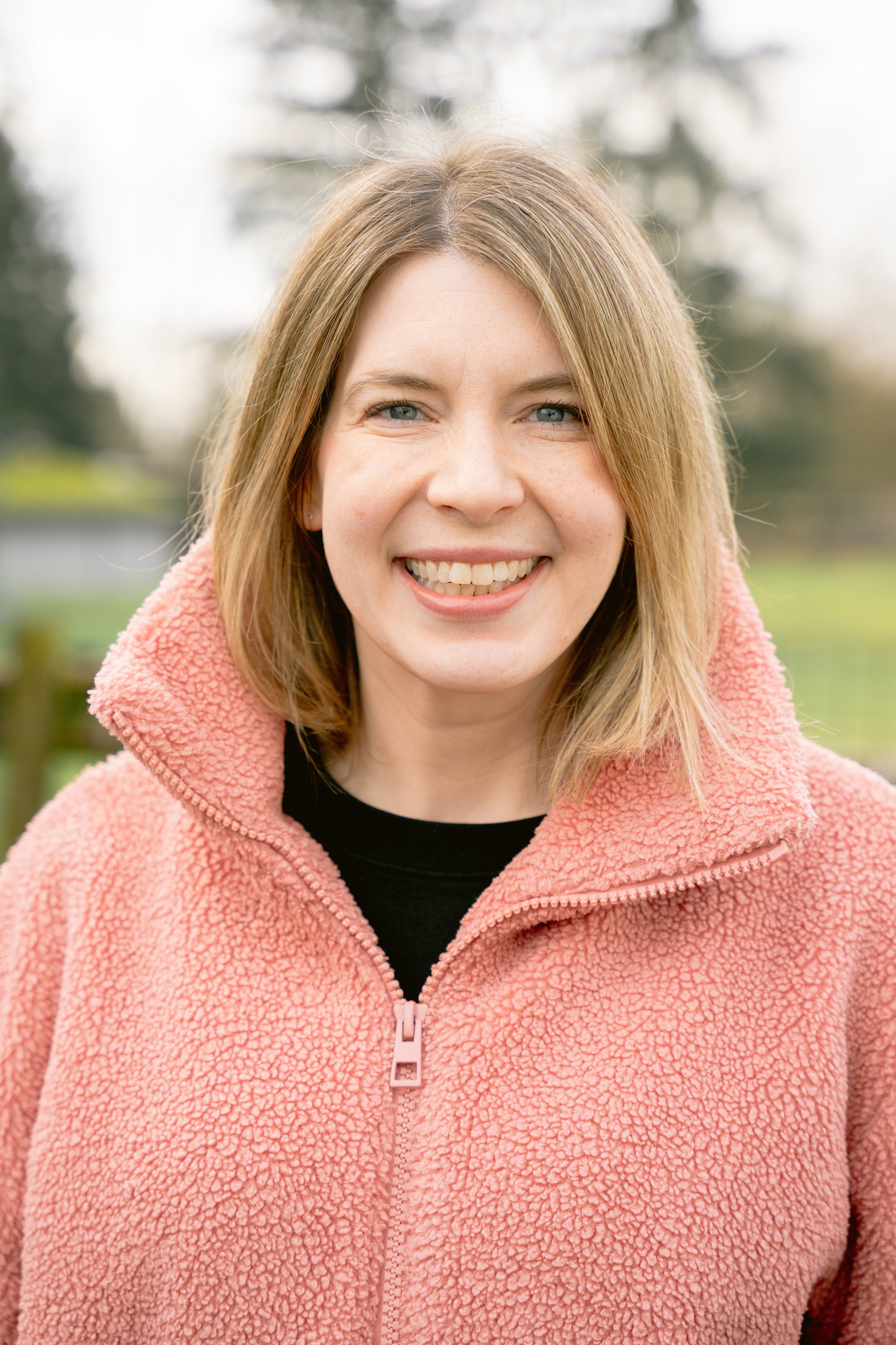A woman with shoulder-length blonde hair and blue eyes smiling outdoors, wearing a pink fleece jacket with a black shirt underneath, with a blurred green and brown outdoor background.