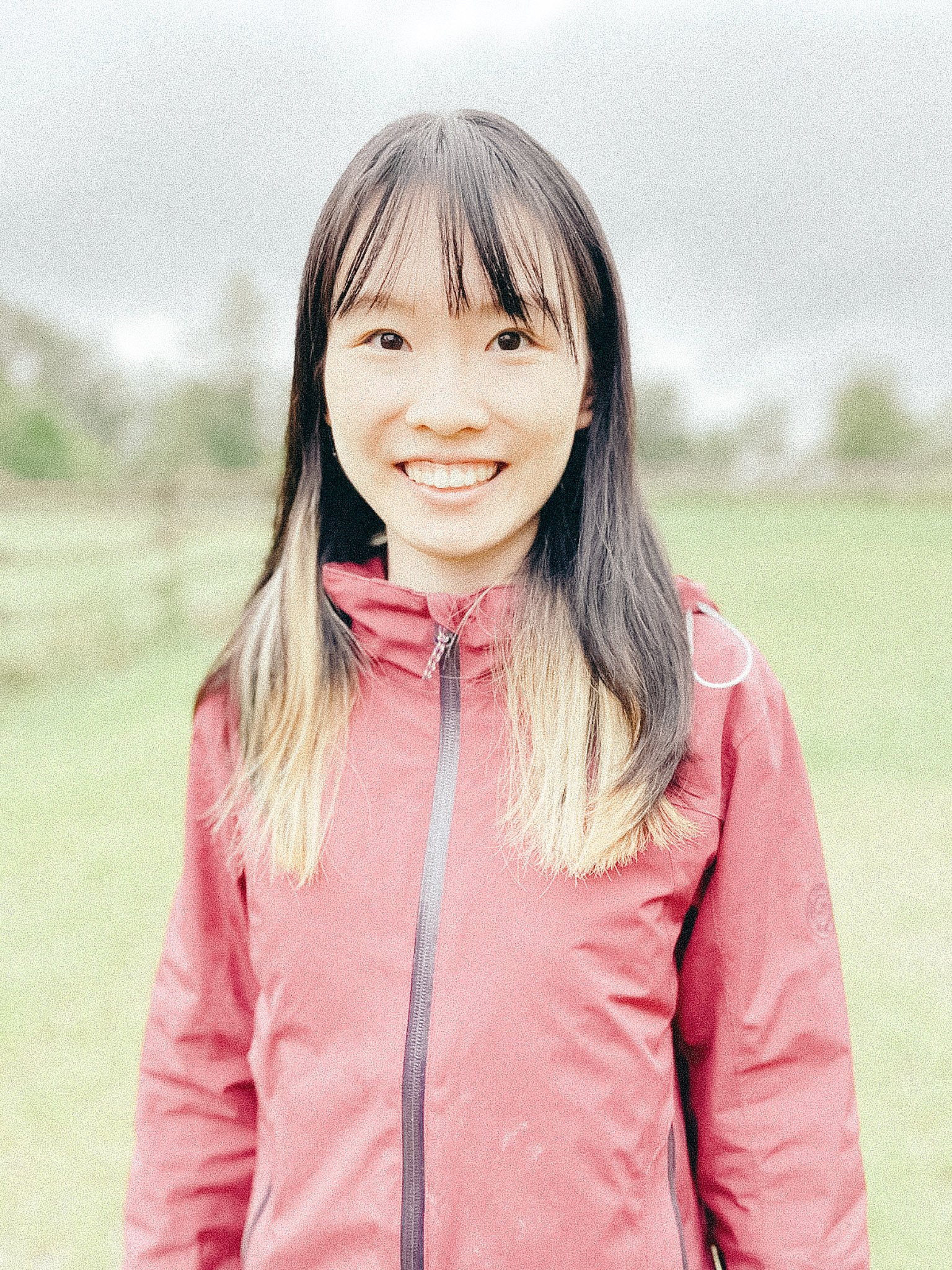 Smiling woman outdoors wearing a red zip-up jacket, with a blurry grassy field and trees in the background.