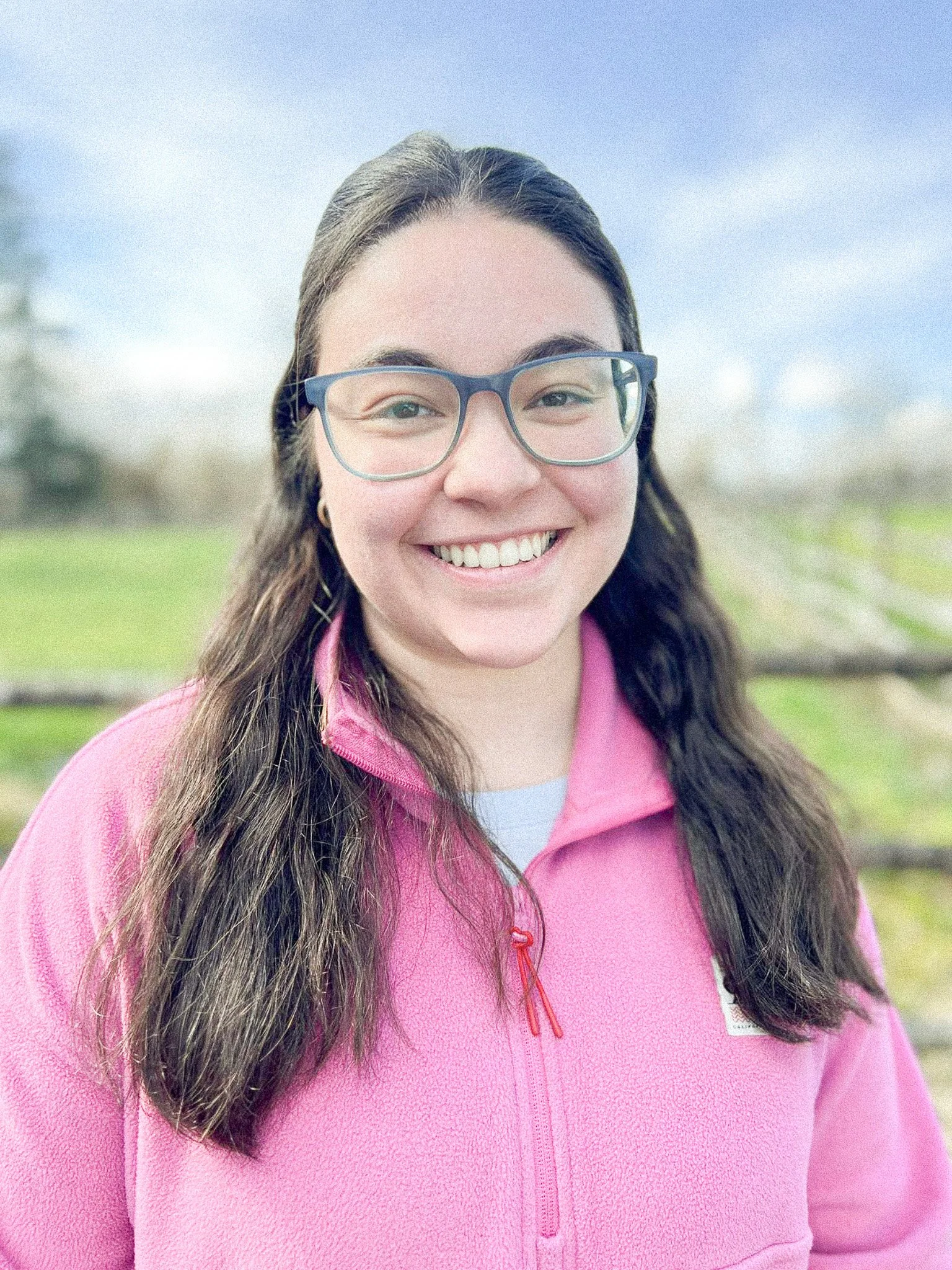 A young woman with glasses smiling outdoors, wearing a pink fleece jacket.