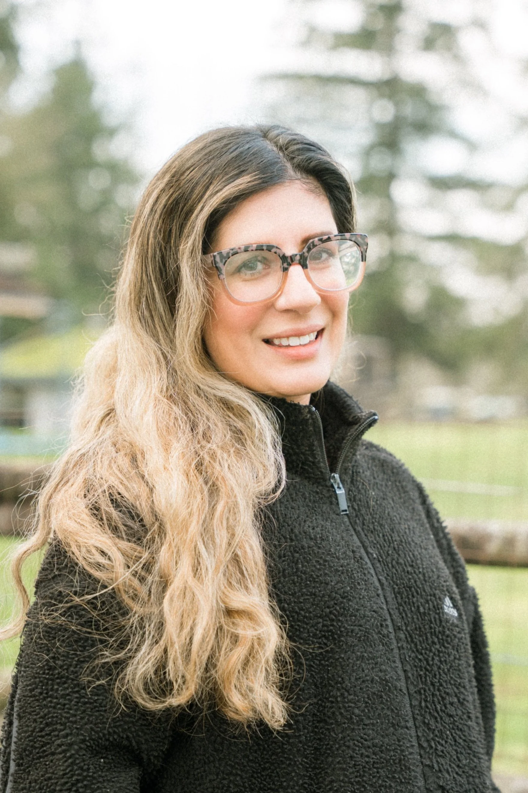 A woman with long, curly blonde hair and glasses smiling outdoors, wearing a black fleece jacket.