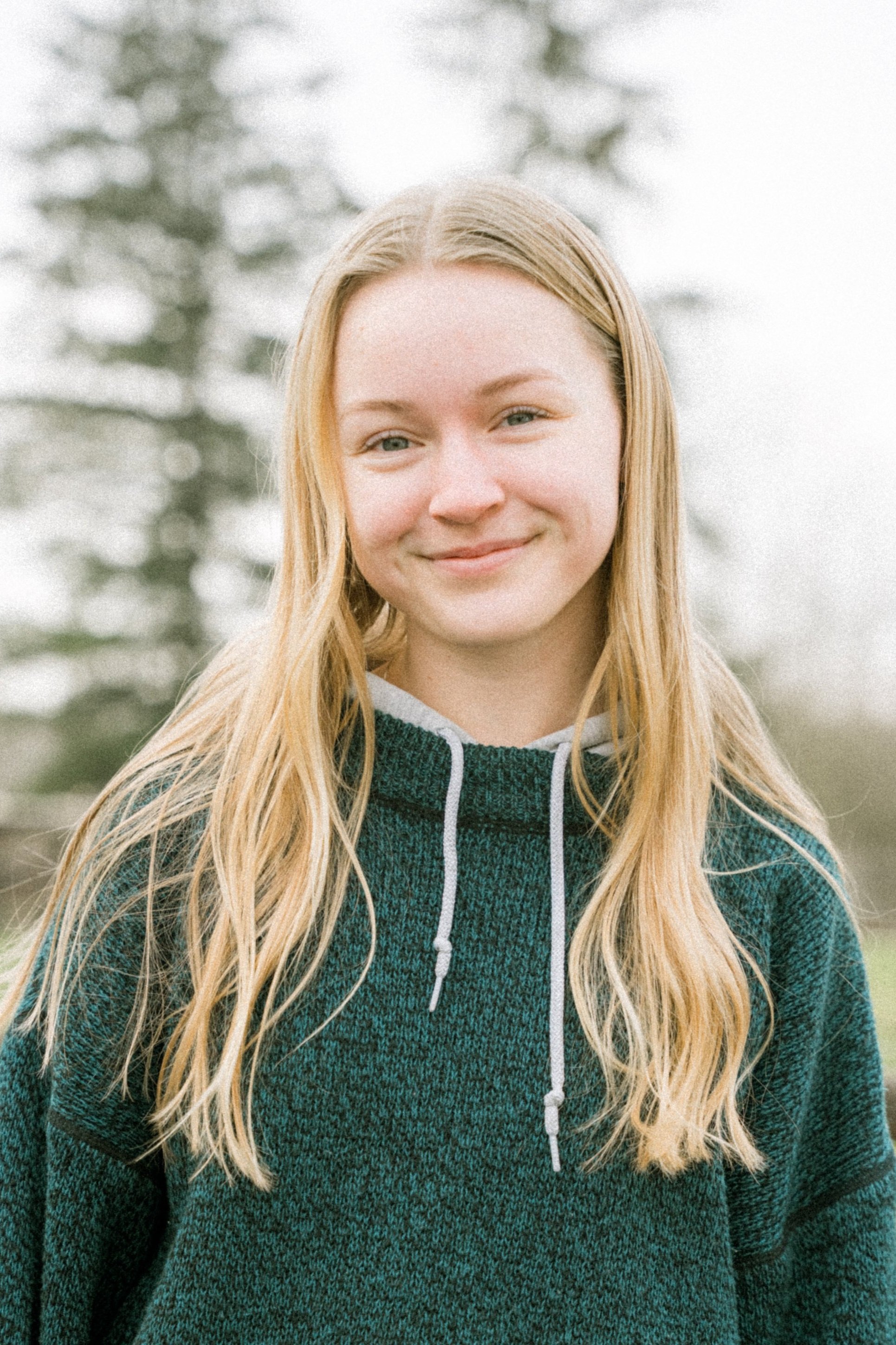 A young woman with long blonde hair smiling outdoors in front of blurred trees, wearing a green and gray hooded sweater.