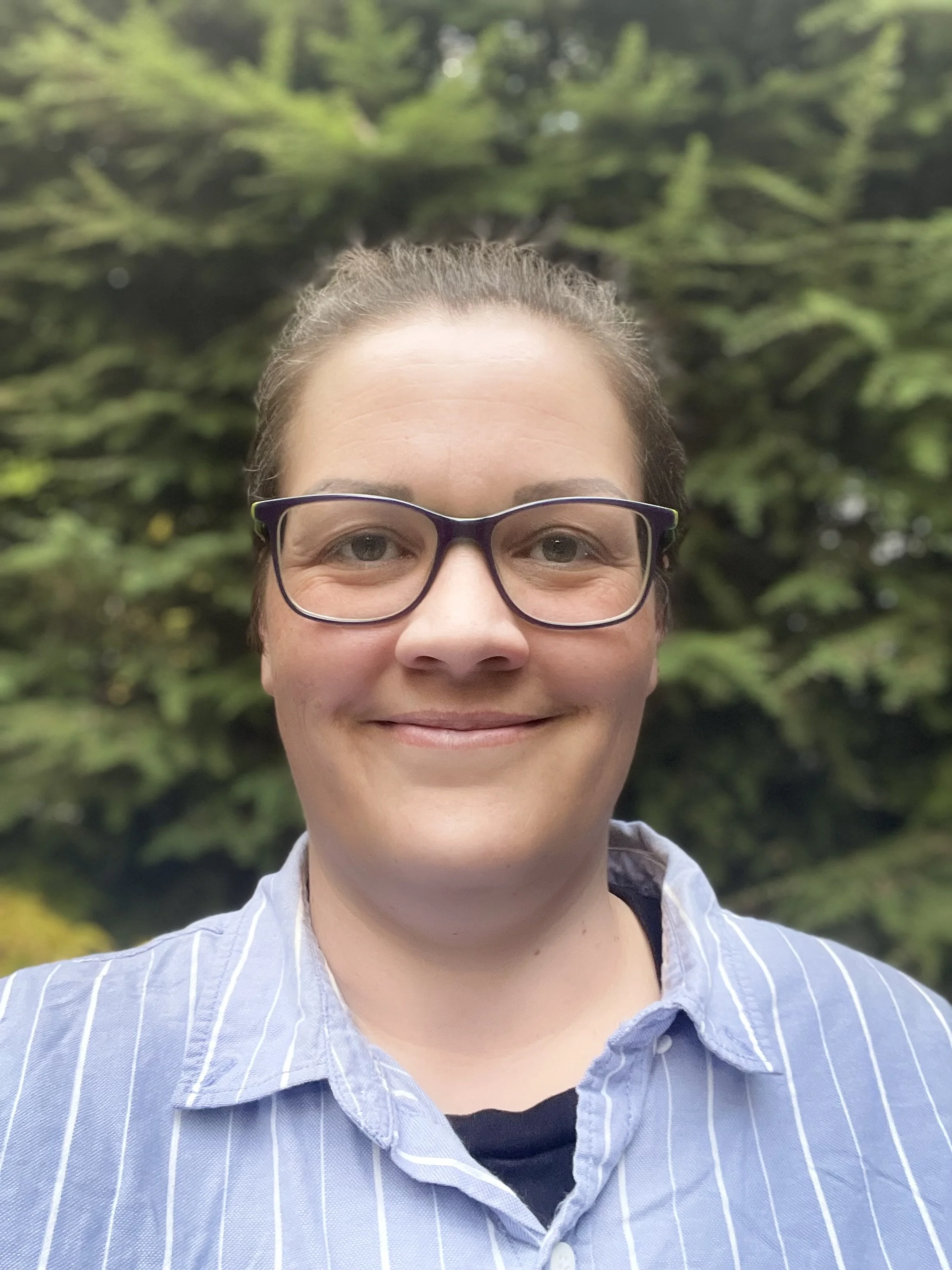A woman smiling outdoors with green trees in the background, wearing glasses and a striped blue and white shirt.
