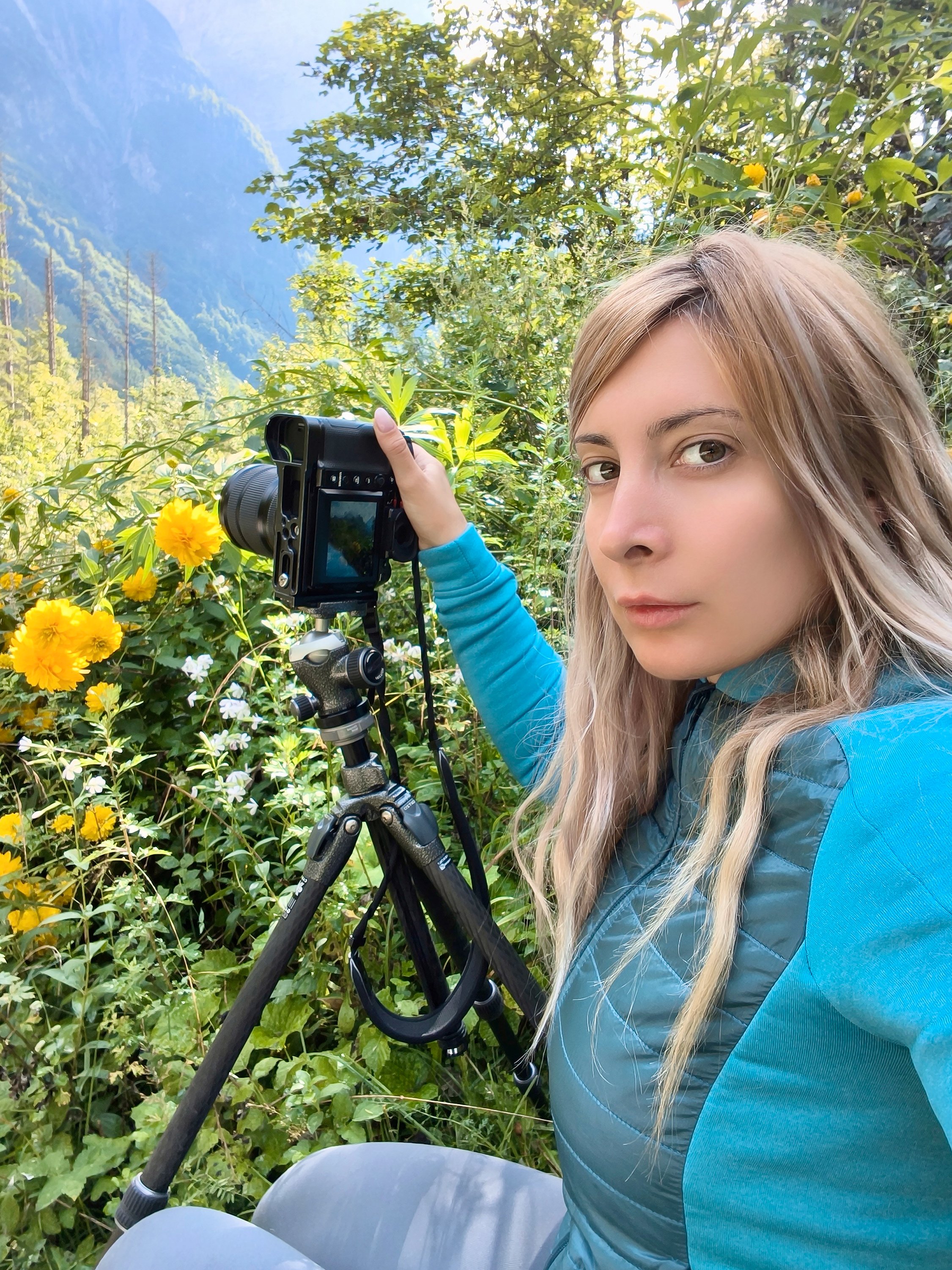 A woman with long blonde hair wearing a blue jacket is outdoors in a lush garden with yellow flowers and green foliage, holding a camera mounted on a tripod.