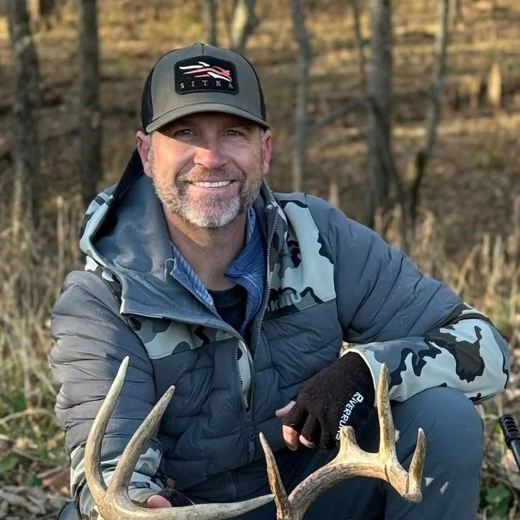 Man in camouflage jacket and black cap holding a deer antler outdoors in a wooded area.