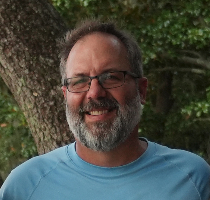 Smiling middle-aged man with glasses and a beard outdoors near a tree, wearing a light blue shirt.