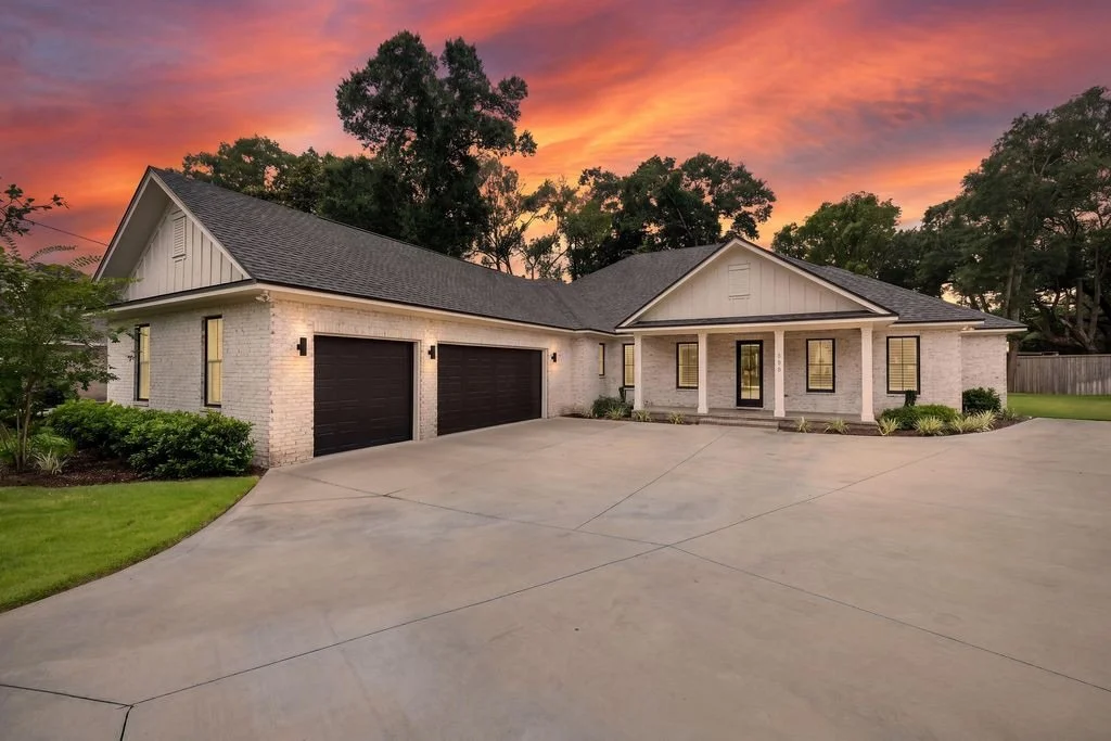 A modern single-story house with a gray brick exterior, black garage doors, and a front porch set against a vibrant sunset sky, surrounded by a well-maintained lawn and landscaping.