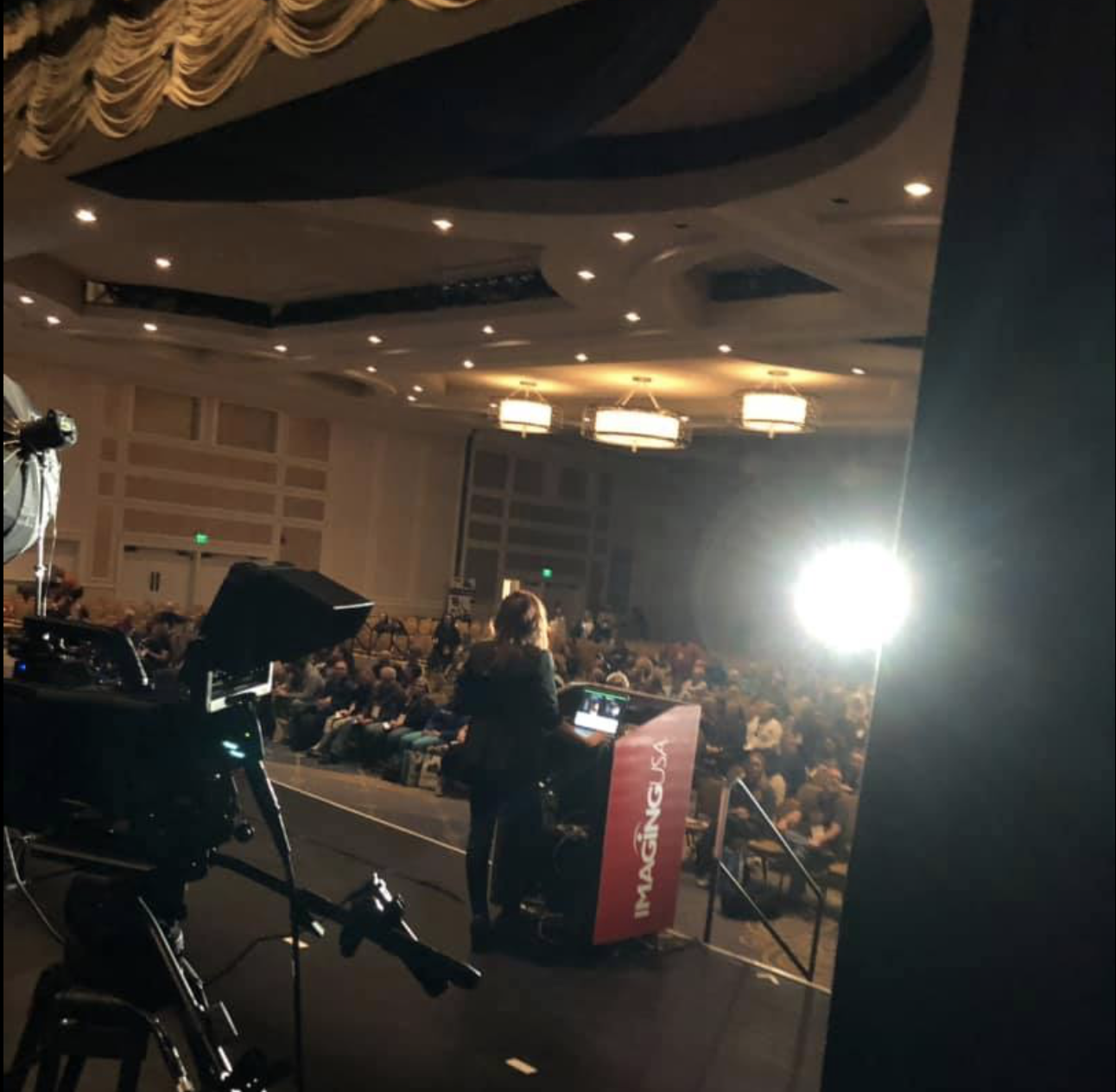 View of a large conference or event hall with many attendees sitting in chairs, a woman standing at a podium labeled 'IMAGING USA', and professional camera equipment capturing the event. Bright stage lights illuminate the scene.