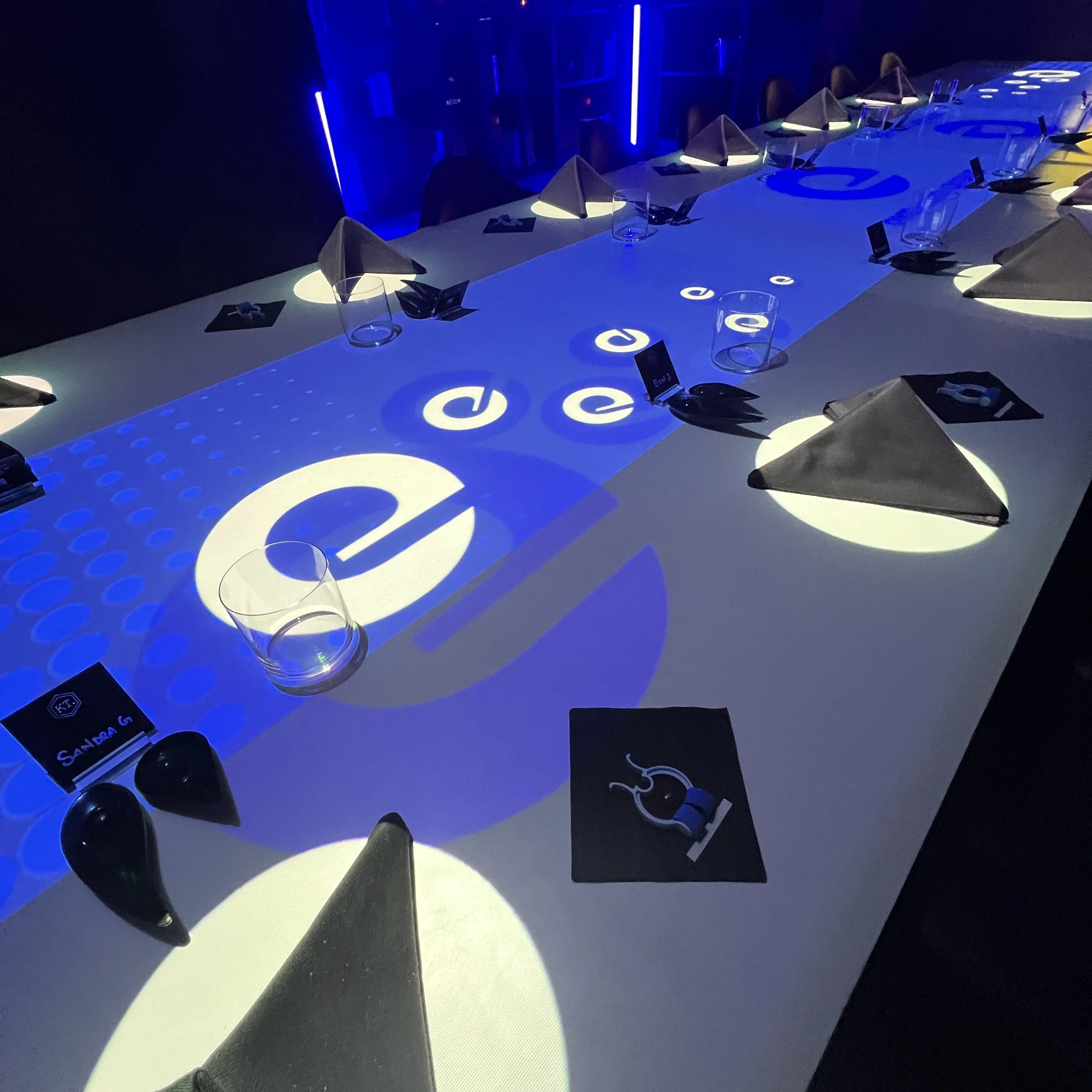A table set with black napkins folded into triangles, empty glasses, and black place cards, illuminated with blue and white lighting projecting the letter 'e' repeatedly across the table surface.