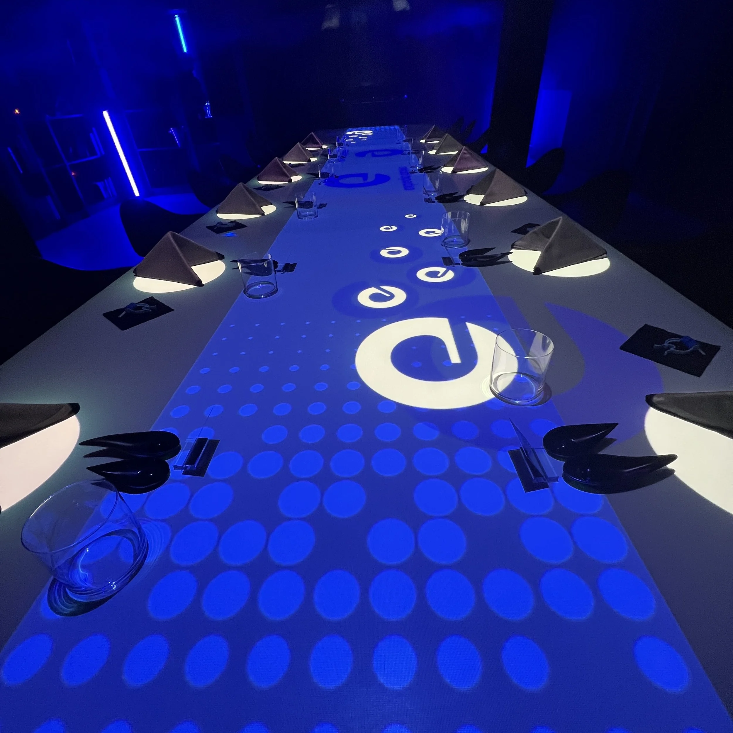 A long conference table illuminated with blue lighting and a projection spelling 'decc' with circular patterns on the surface, set with glassware, black napkins, and small cards.