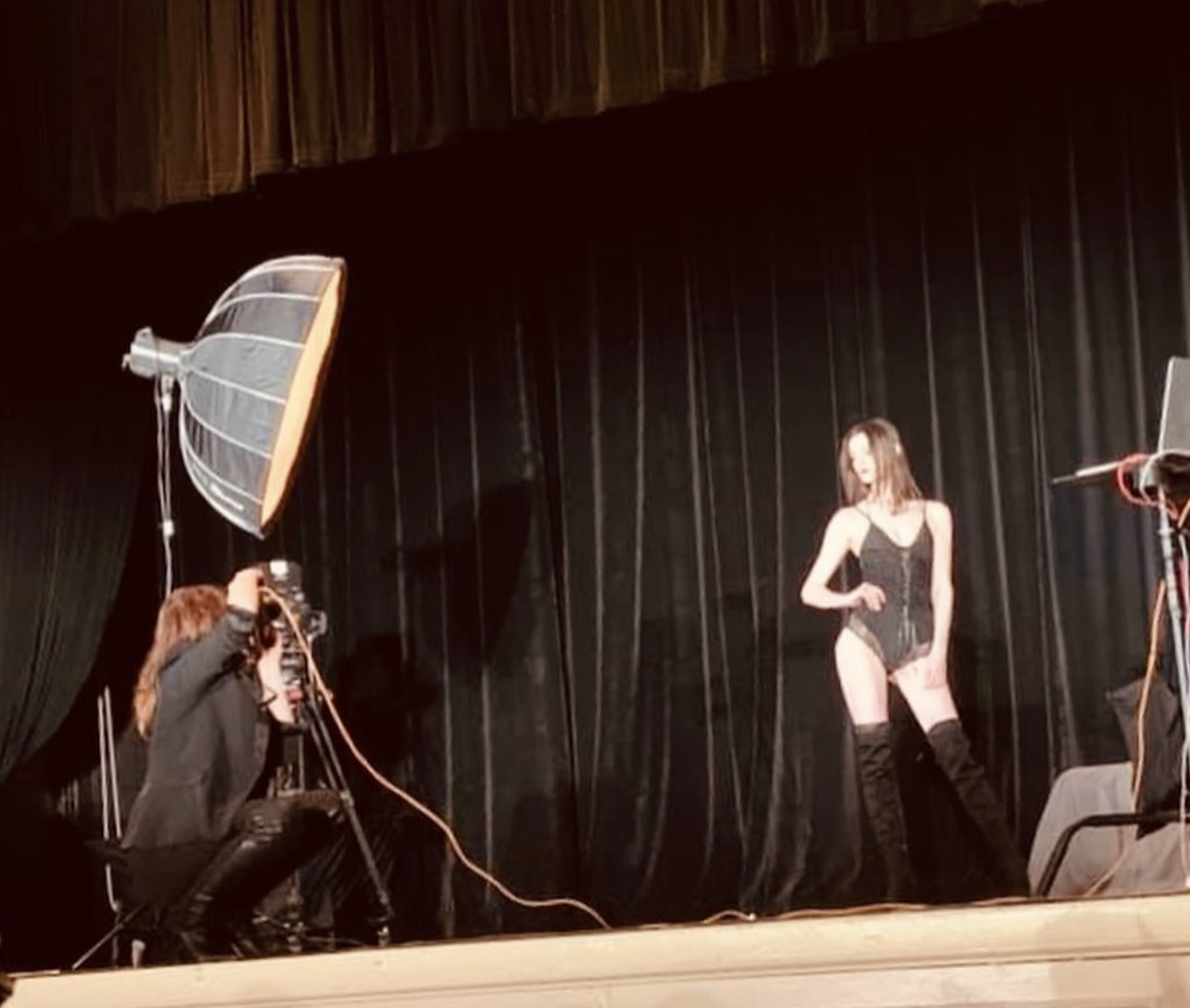 A woman in black lingerie and thigh-high boots strikes a pose on a stage, with a photographer capturing her picture, and studio lights illuminating the scene.
