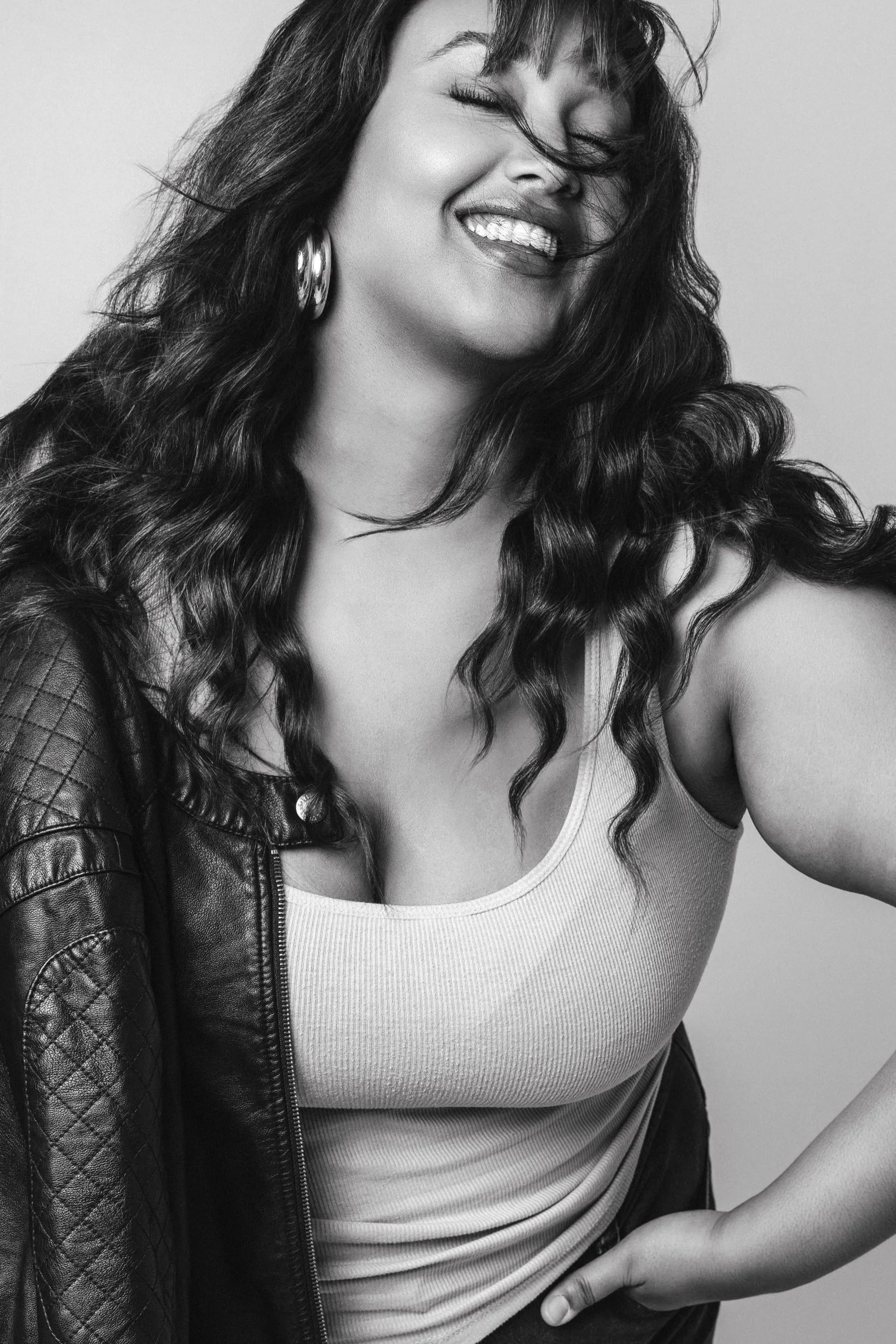 Black and white photo of a woman with long, curly hair, smiling and winking, wearing a tank top, earrings, and a leather jacket.