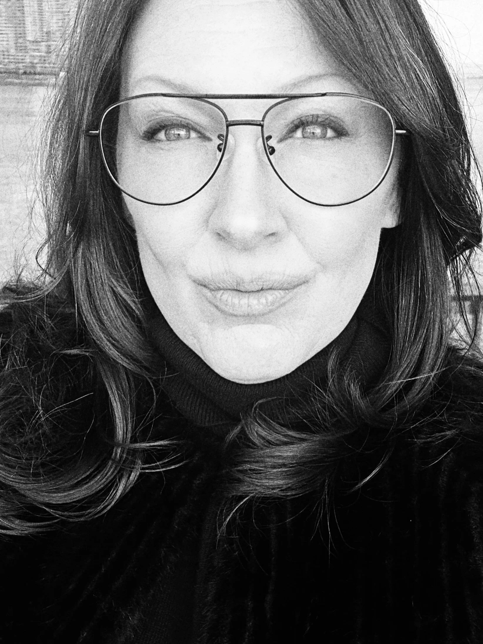 A woman with long dark hair and large glasses smiling, wearing a dark coat or sweater.