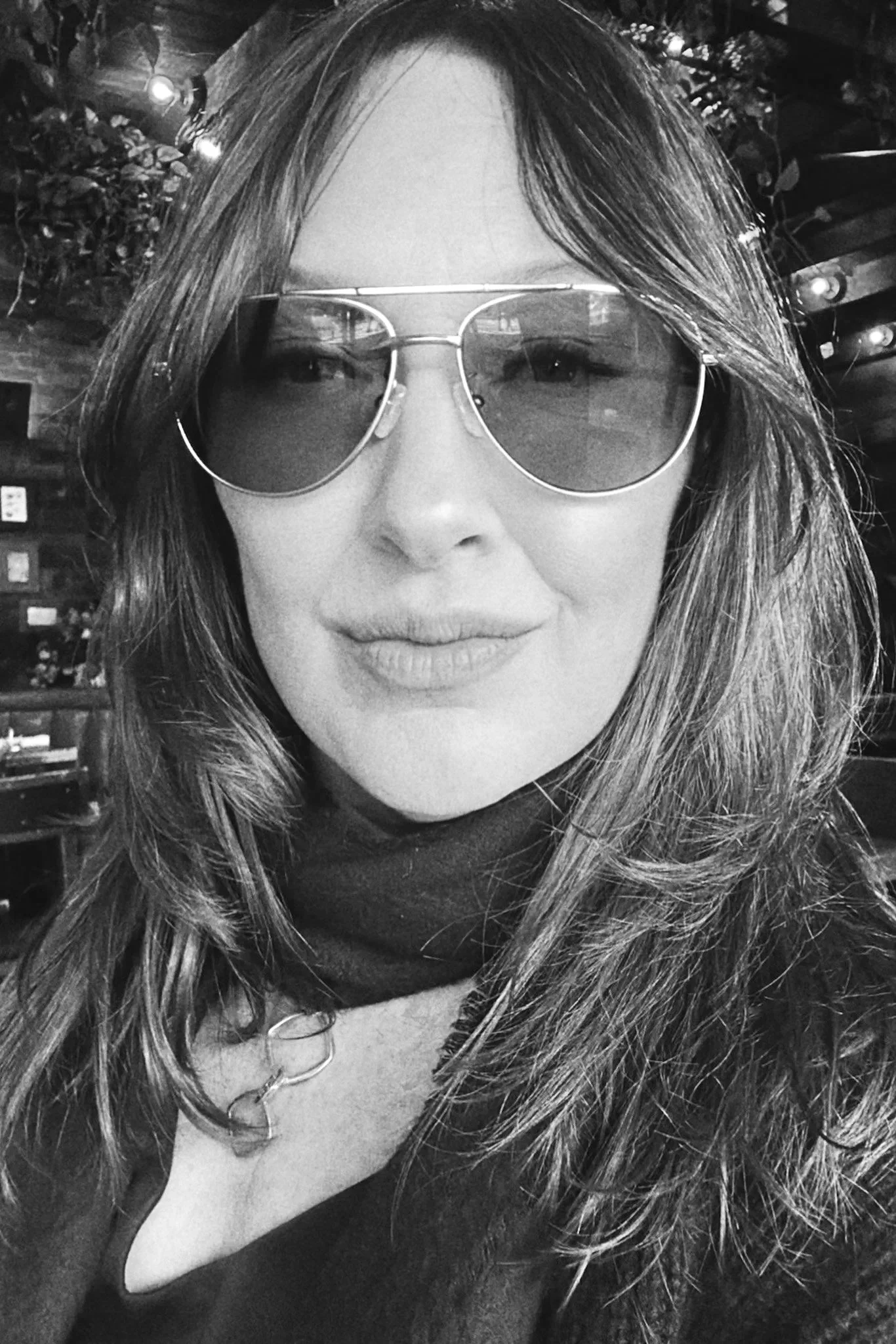 A woman wearing sunglasses and a dark neck scarf in a dimly lit indoor setting.