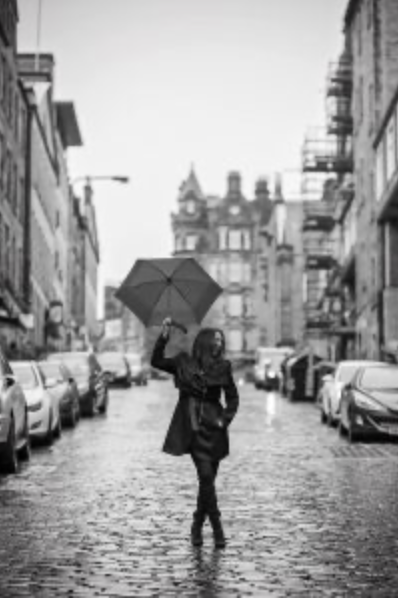 Person holding an umbrella walking on a wet city street, lined with parked cars and historic buildings.