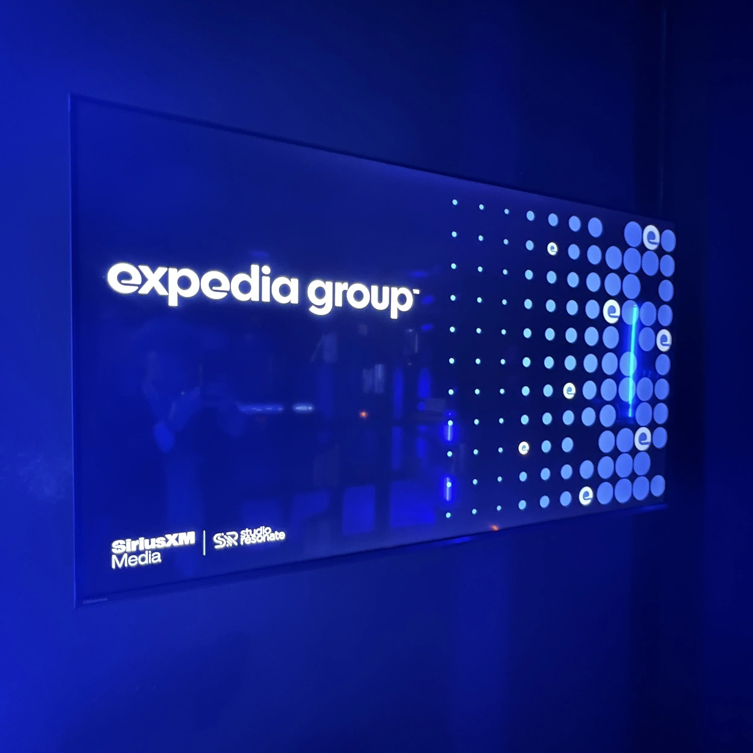 Photo of an illuminated blue sign with 'Expedia Group' logo, SiriusXM Media and Studio Resonate logos below, reflected on a glossy surface.