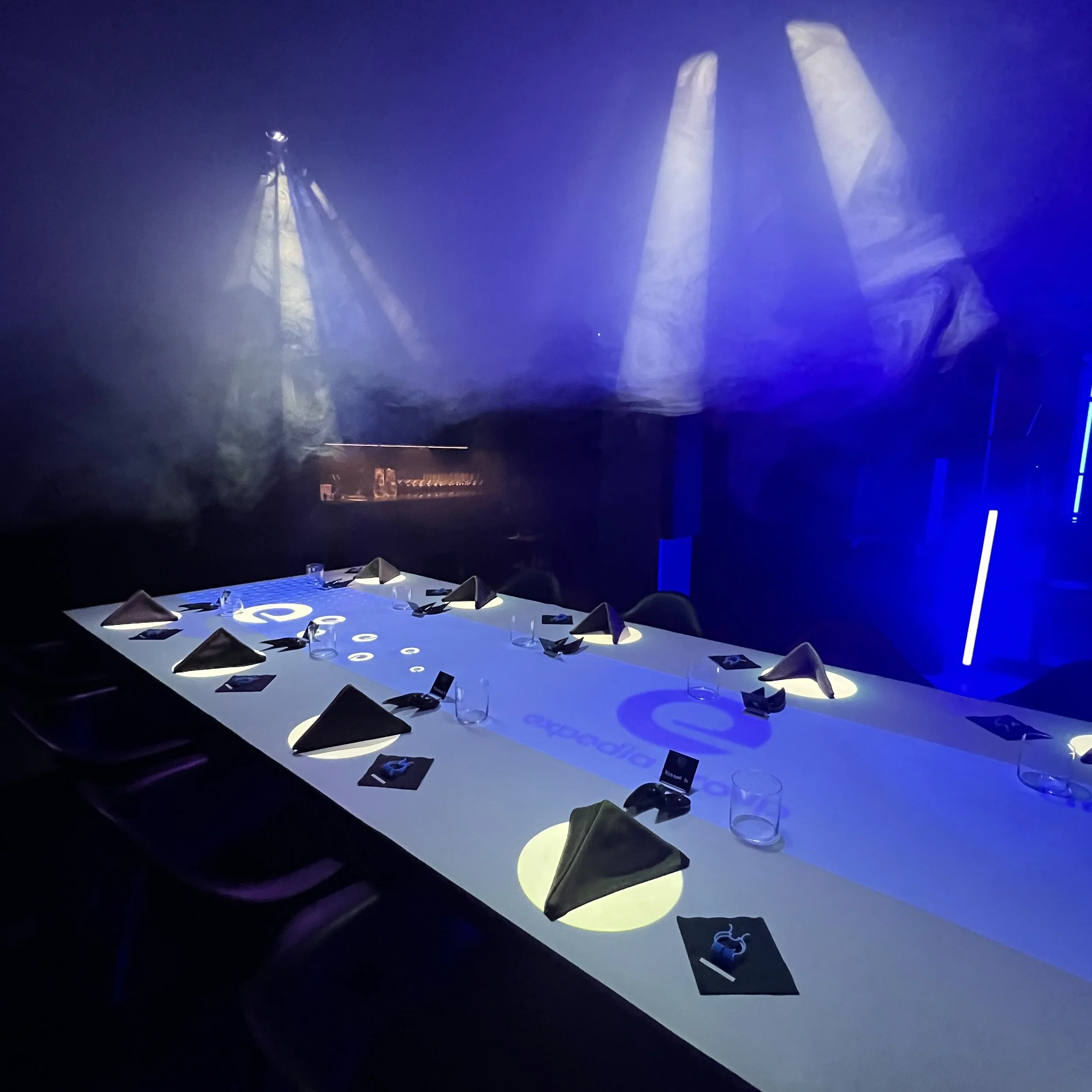 A dimly lit conference room with a long table set with napkins, glasses, and small informational cards, illuminated by blue and purple lighting with beams overhead.