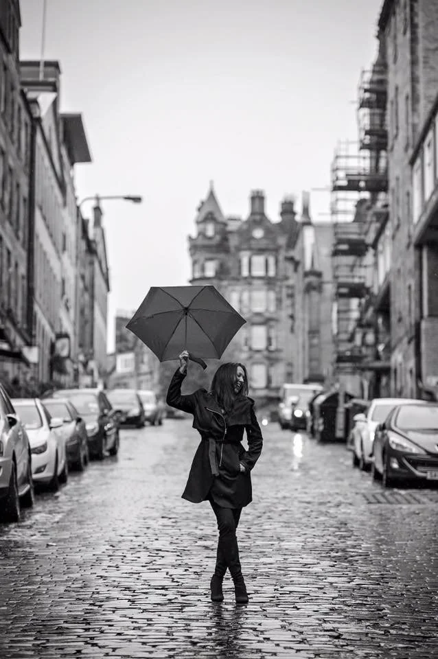 A woman walking on a wet cobblestone street in a city holding an umbrella, with buildings and parked cars lining the street.