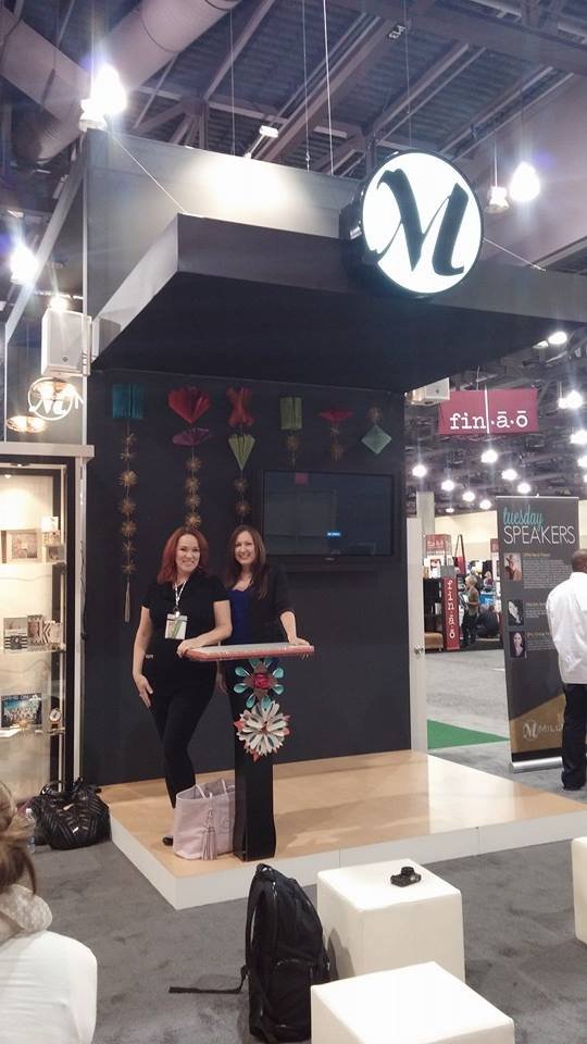 Two women standing on a small stage at a booth with a black backdrop and a large white circle sign with a black letter 'N'. The women are smiling and standing near a small table decorated with a floral pattern. The booth is part of a trade show or ex
