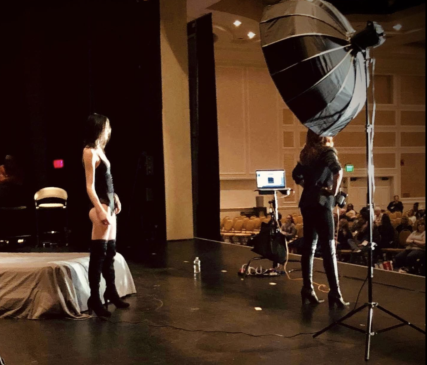 A woman in a black outfit and high boots stands on a stage with her back to a large studio light, preparing for a photo or performance. Audience members are seated in the background.
