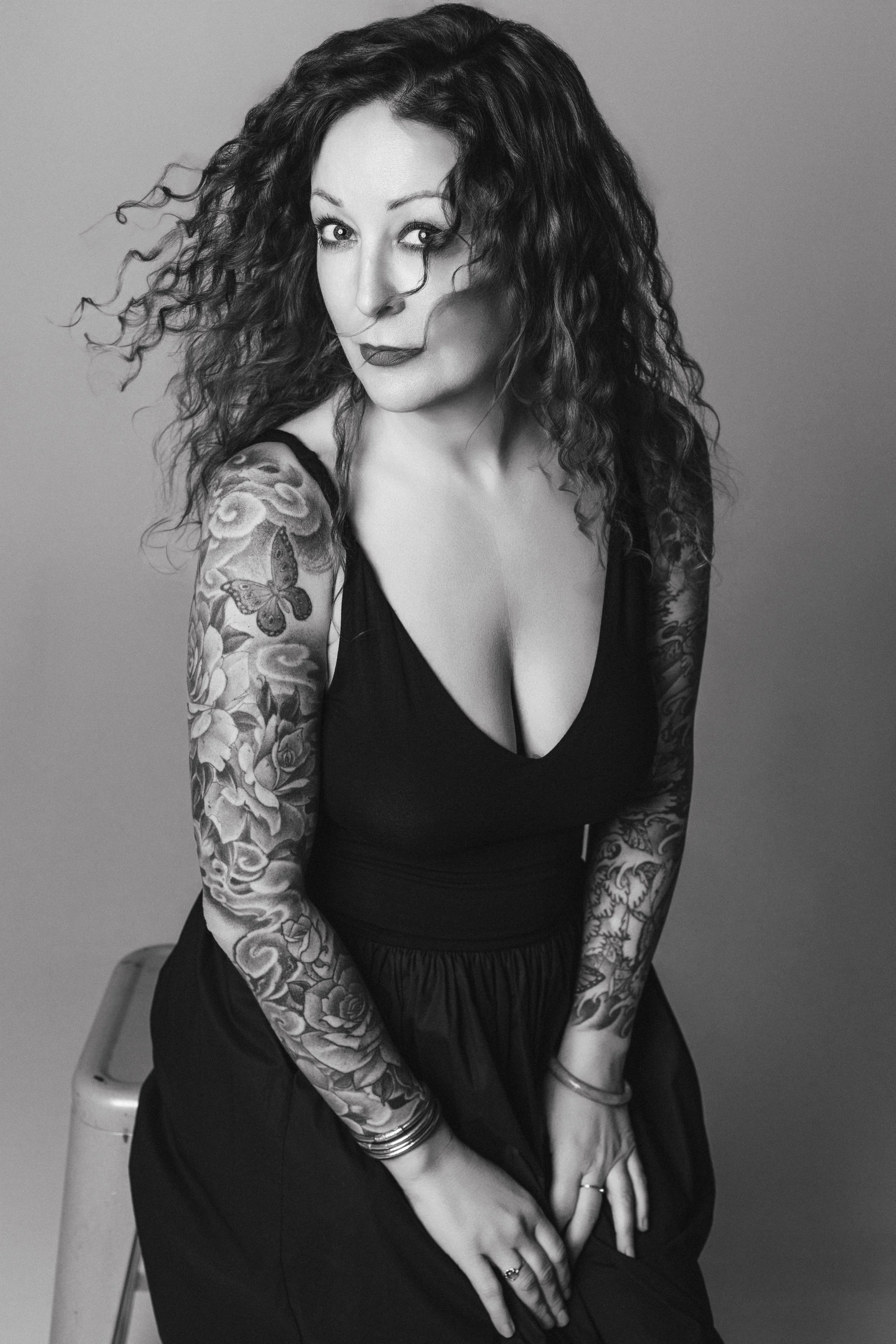 A woman with curly hair and tattoos on her arms, wearing a low-cut black dress, sitting on a stool against a plain background.