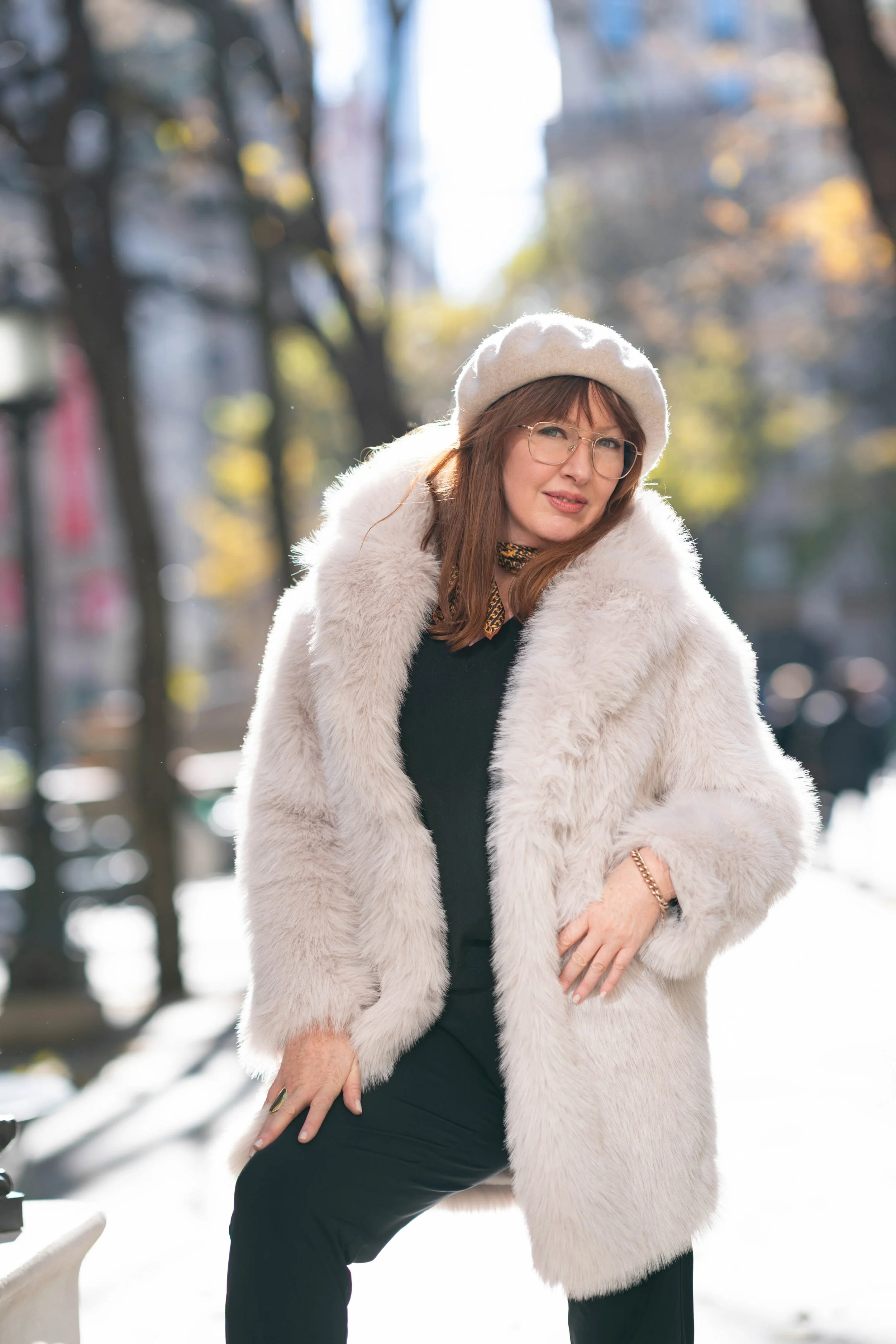A woman wearing a white fur coat, white beret, and glasses standing outdoors on a city street with trees and buildings in the background.
