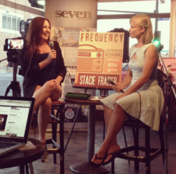 Two women sit on chairs in a radio station or interview setting, with a sign that reads "Frequency" and "Stacie Frasier". One woman with red hair holds a microphone, while the other woman with blonde hair listens. There is a laptop and camera recordi
