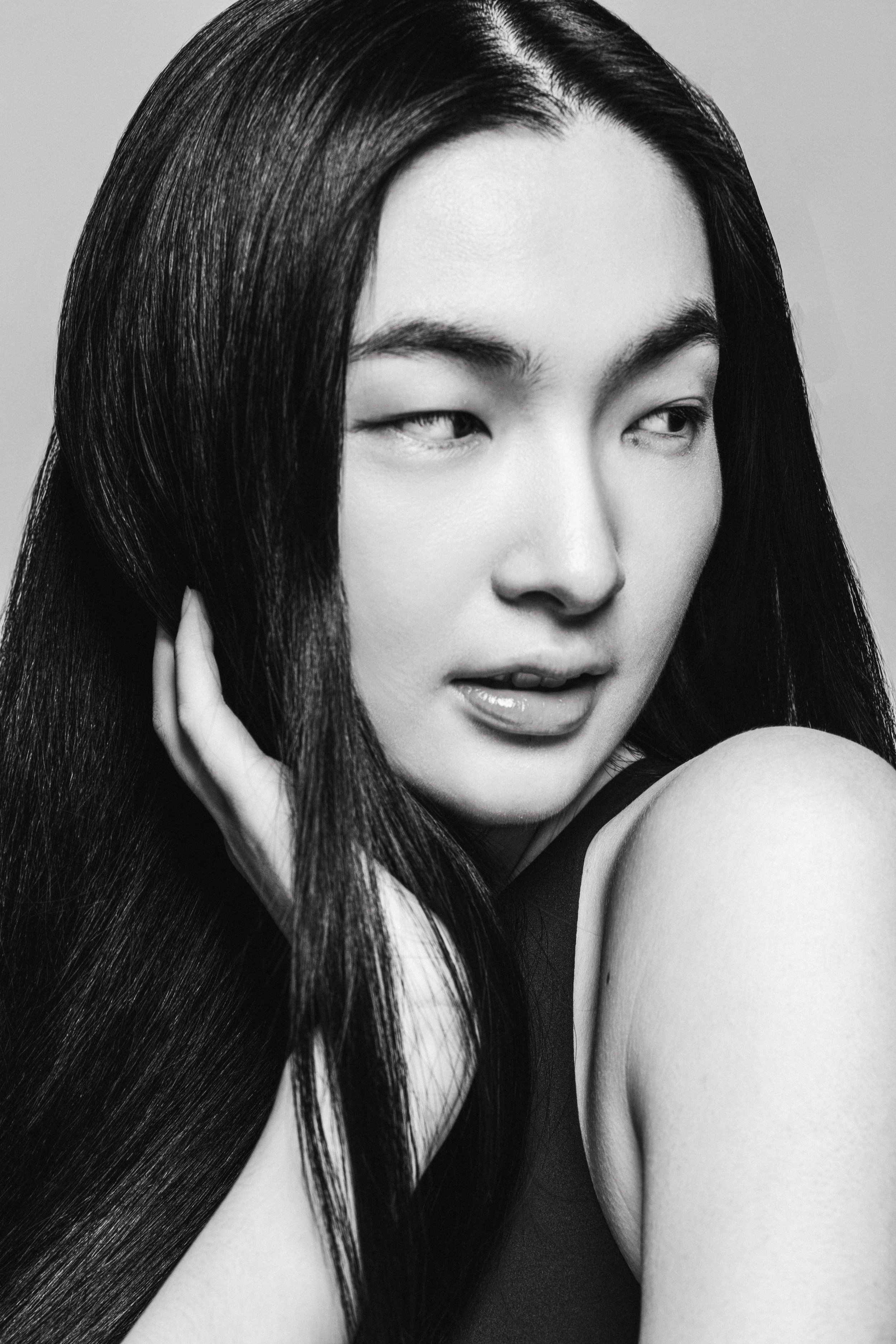 Black and white close-up portrait of a woman with long, straight hair, one eye partially closed, and a hand gently touching her face.