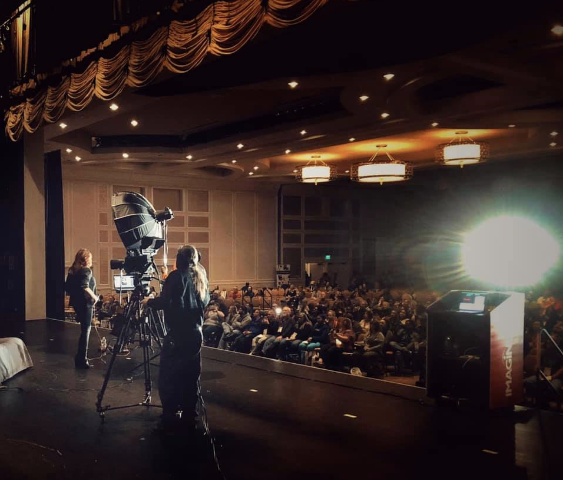 People preparing for a presentation on a stage in an auditorium, with an Audience seated and a bright light shining on the stage.