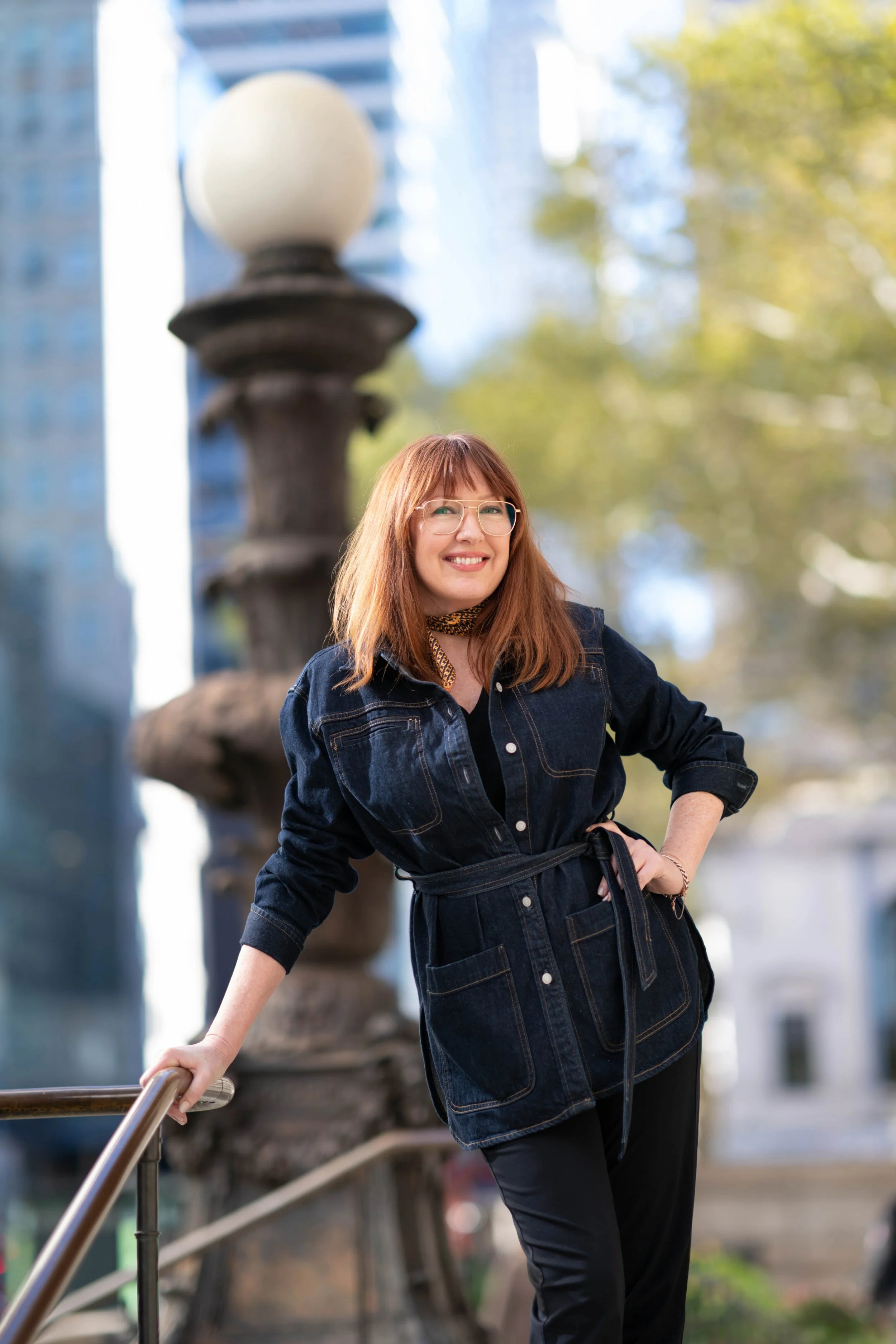 A smiling woman with red hair and glasses standing outdoors, leaning on a metal handrail with one hand, in front of a fountain and urban buildings, on a sunny day.
