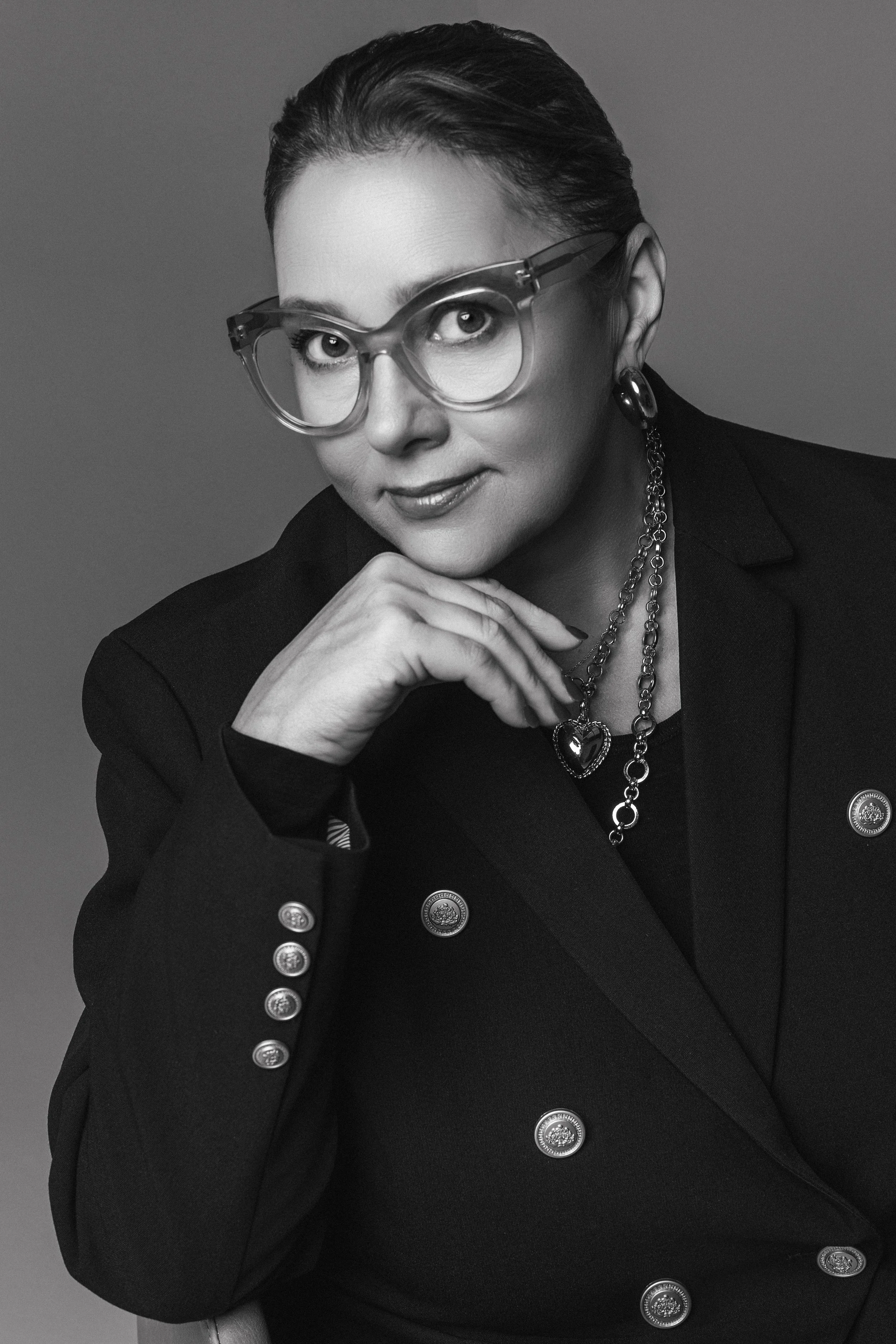 Black and white portrait of a woman wearing glasses, a dark blazer with embellishments, and jewelry including a heart-shaped pendant necklace and hoop earrings, resting her chin on her hand.
