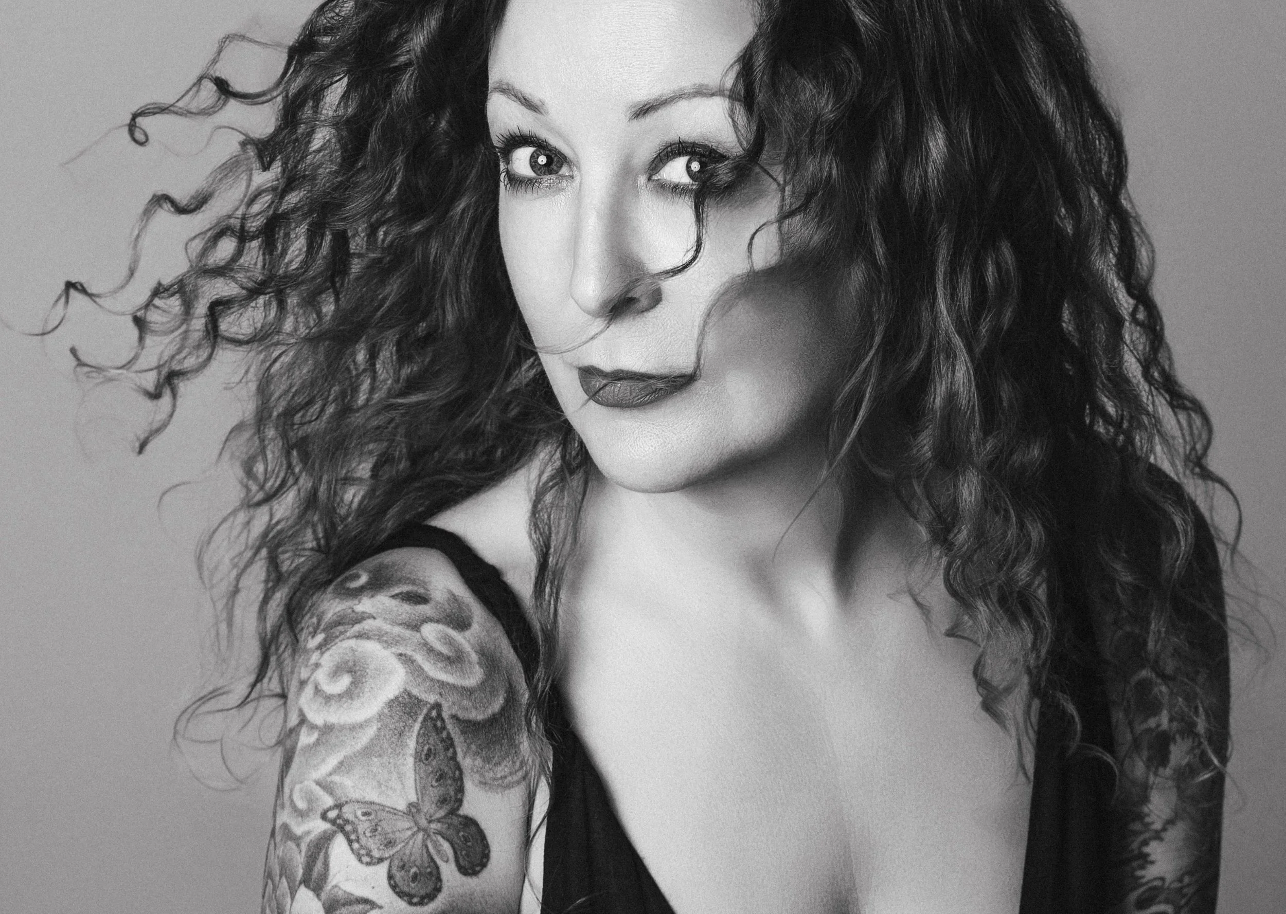A black and white portrait of a woman with curly hair, wearing a sleeveless top, showing a tattoo of a butterfly and roses on her upper arm.
