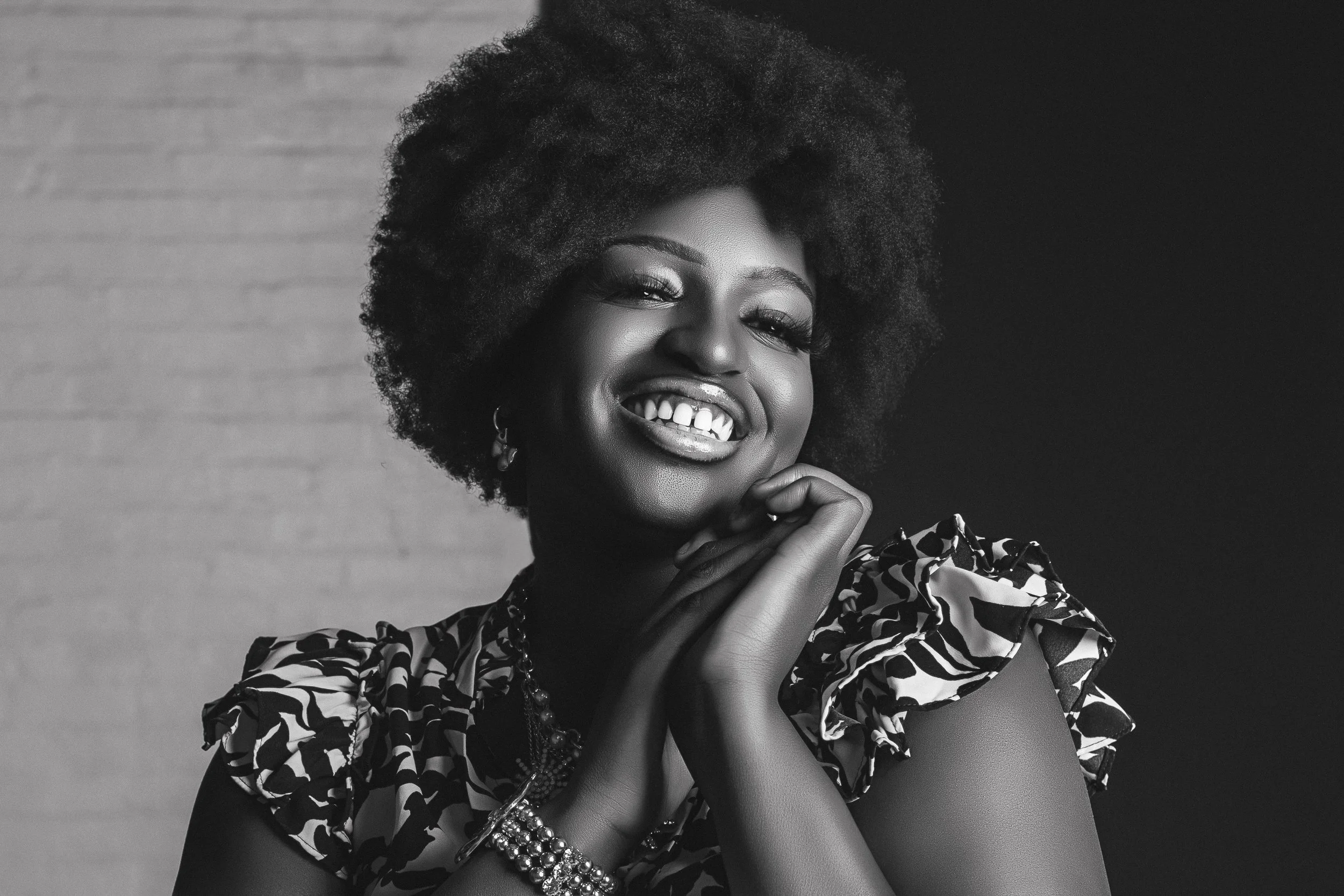 A smiling Black woman with a large afro hairstyle, wearing a patterned dress with puffed sleeves, jewelry, and showing her teeth in a playful expression on a randomly divided background.