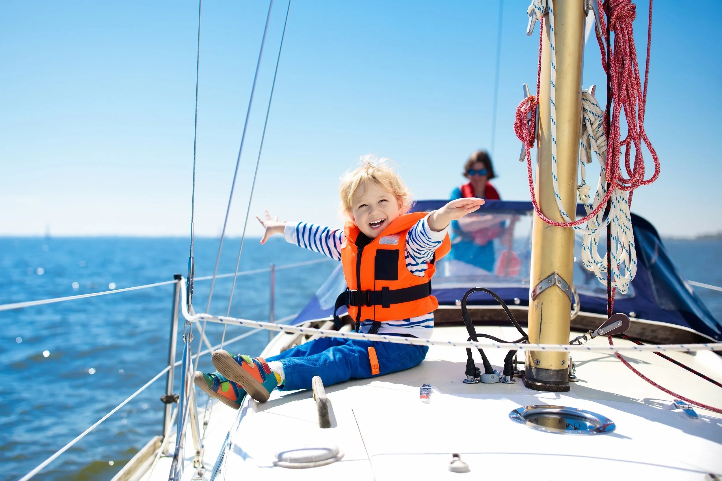 Child wearing an orange life jacket sitting on a sailboat with outstretched arms, blue ocean and sky background.
