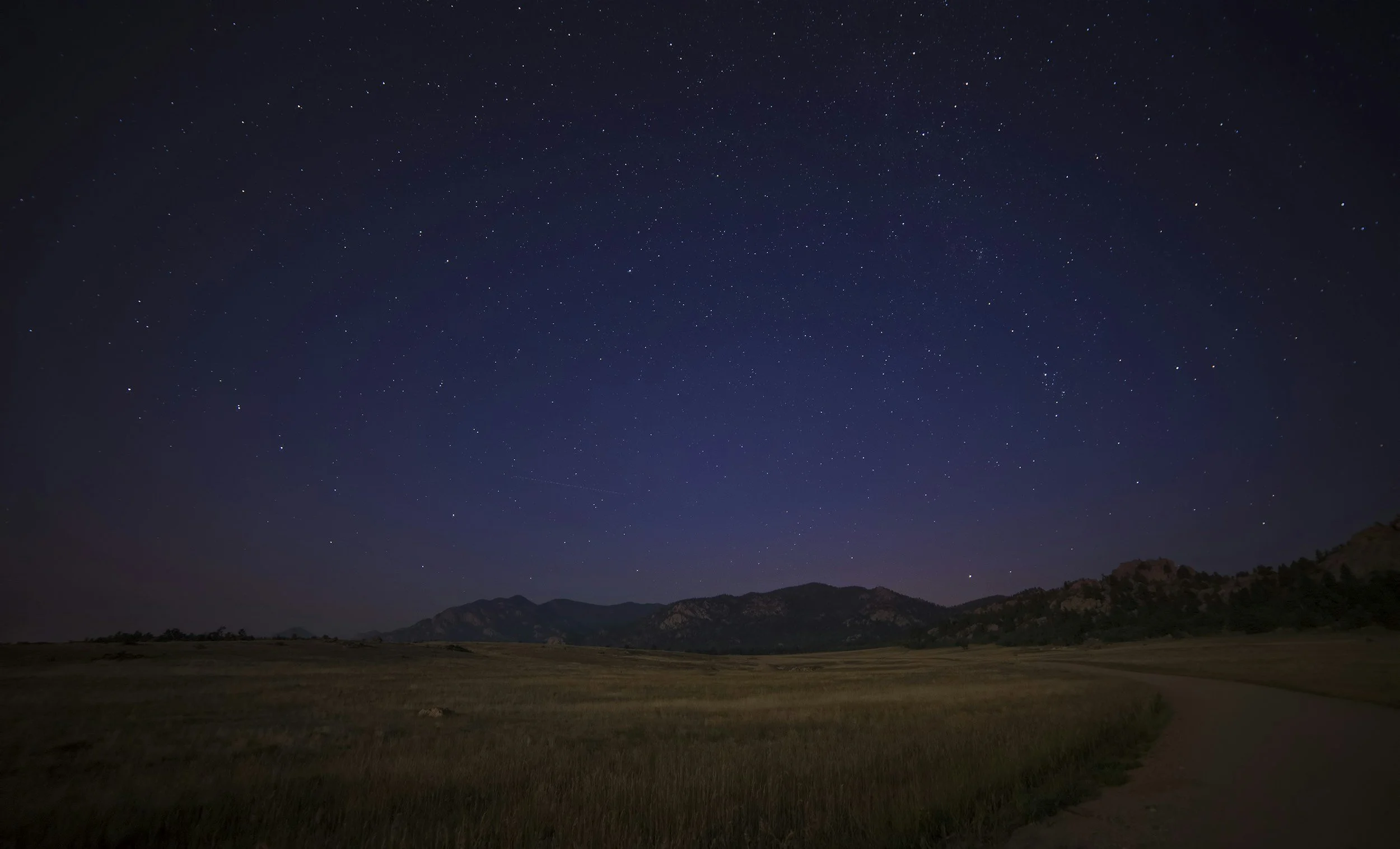 Nighttime landscape with a clear starry sky above open fields and distant mountains.