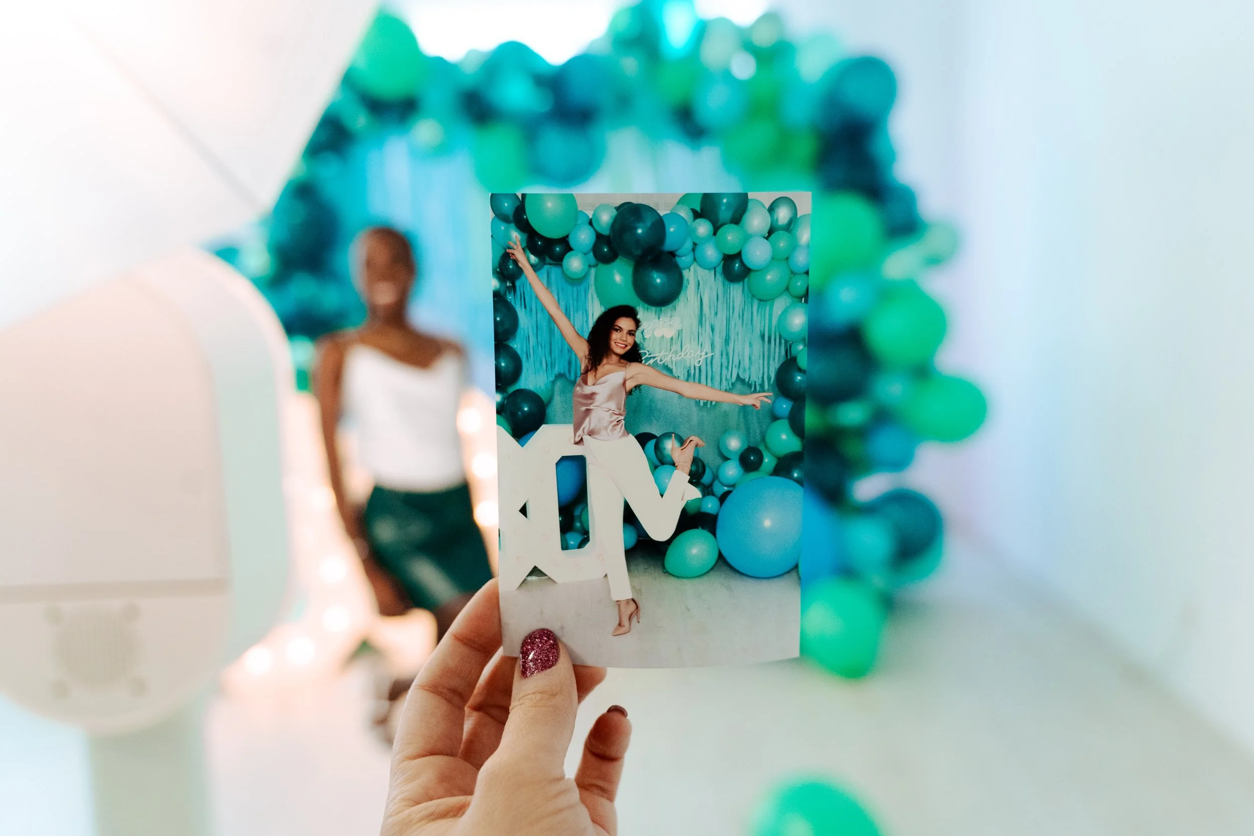Photo of a hand in the foreground holding a photo of a woman posing happily.  In the background there is a separate woman smiling but blurred out.