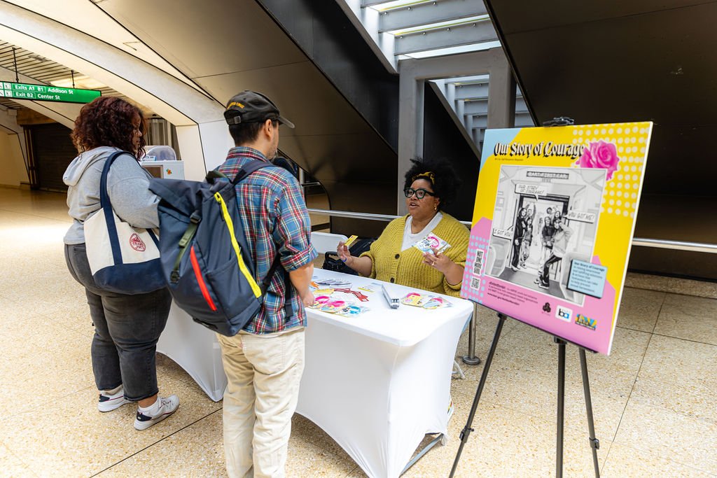  Two people in front of a white table talking to someone seated and promoting the Our Story of Courage campaign 