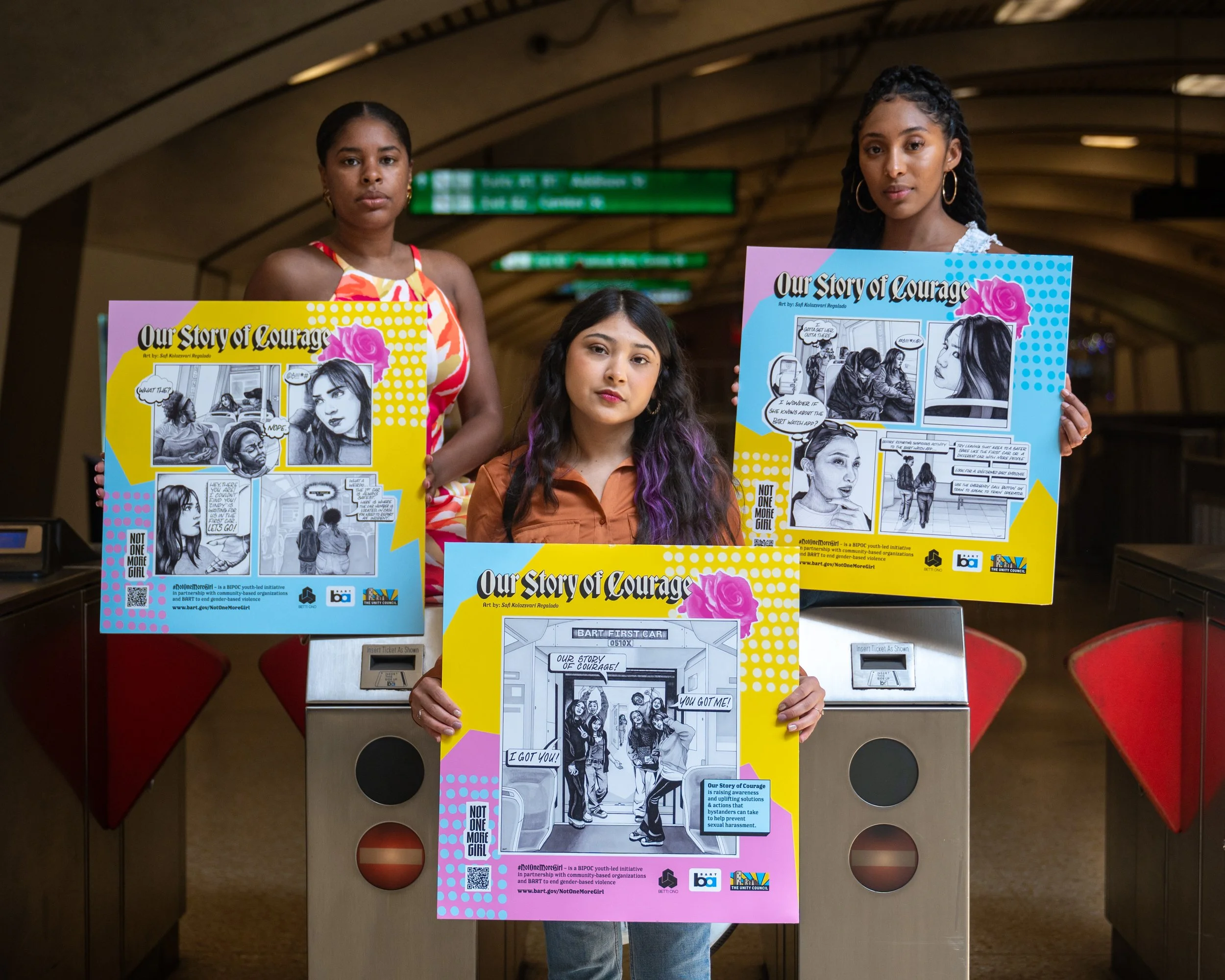 Three women holding posters of the Our Story of Courage campaign inside a BART station