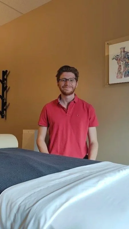 A young man with glasses and a beard wearing a red polo shirt, standing in a hotel room with beige walls, a framed colorful art piece, and a bed with white sheets.